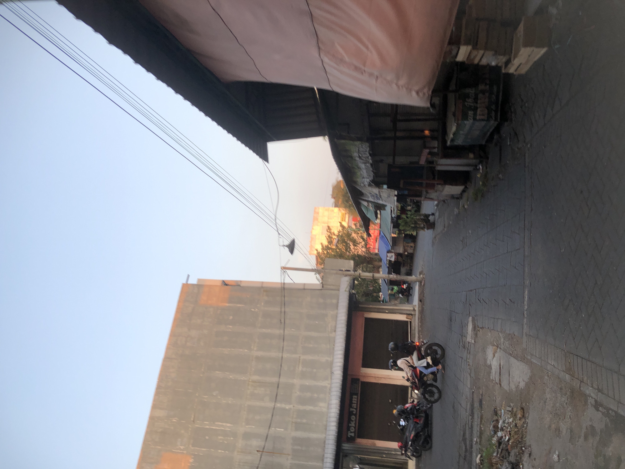 The image depicts a street scene in Sidoarjo, Indonesia, likely at dusk or dawn given the soft lighting and clear sky. On the left, a large, unfinished or dilapidated concrete building with a light brown facade occupies a significant portion of the frame. Below this building, a storefront with the signage "Toko Jam" is visible, indicating a watch shop. Several motorcycles are parked in front of the shop. Two people are riding a motorcycle down the street, with the rider wearing a light-colored jacket and the passenger in a dark jacket.
To the right of the street, a series of small market stalls or shops are set up under lean-to roofs. One stall has a large, draped pink covering, and another displays wooden crates. The street itself is paved with interlocking gray bricks. The overall impression is of a developing urban area or a market district, with a mix of permanent structures and temporary stalls. The presence of motorcycles and people suggests ongoing activity.