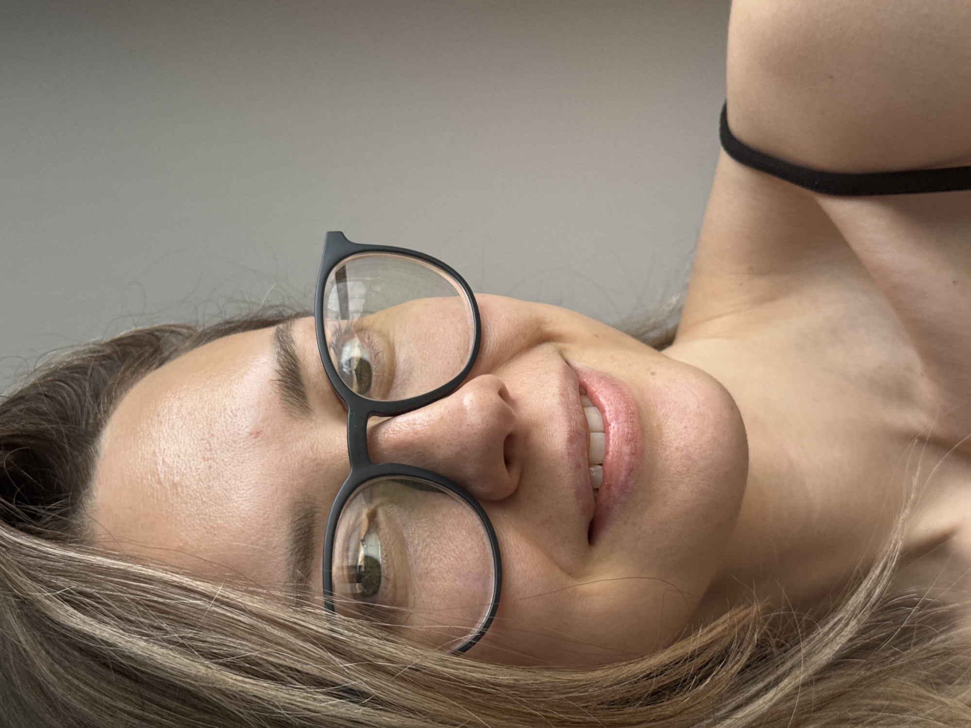 A woman with fair skin, light brown hair, and black-rimmed glasses is lying down. Her head is tilted, and she is looking up. She is wearing a black strap on her shoulder. The background is a solid, light gray color. There is no information in the image or provided to indicate the location is Chaikovsky, Russia.