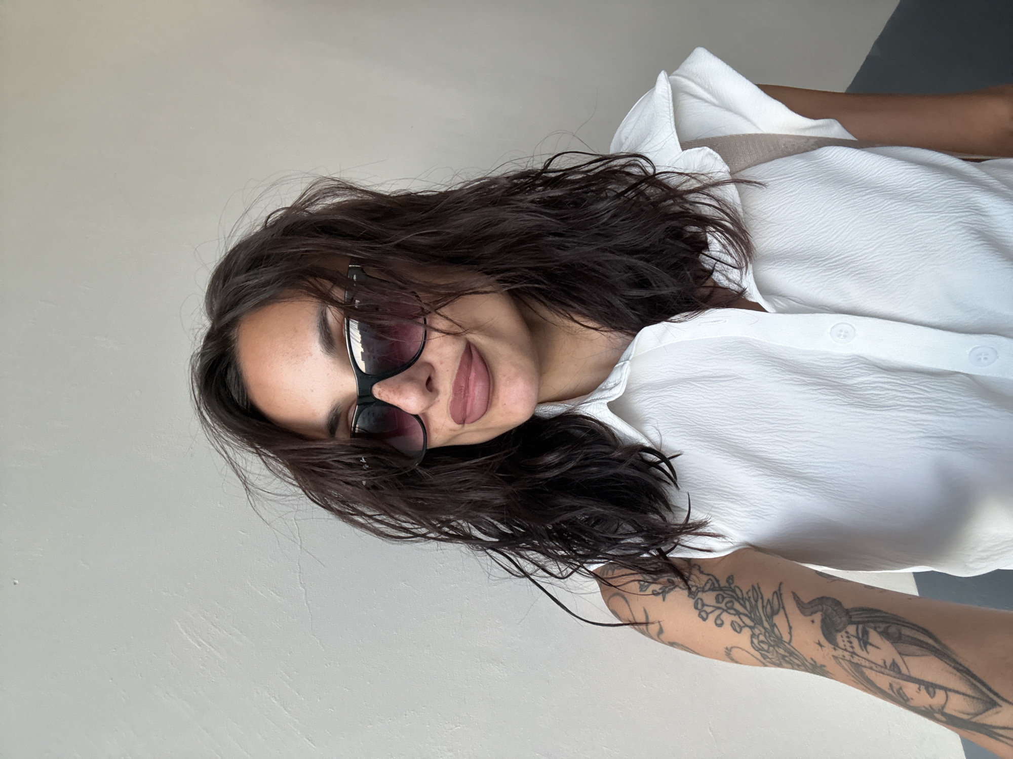 A woman with dark, wavy hair and sunglasses is pictured. She is wearing a white, textured collared shirt and has tattoos on her left arm. She is standing against a plain, textured wall. The location is the city of Pleven, Bulgaria.
