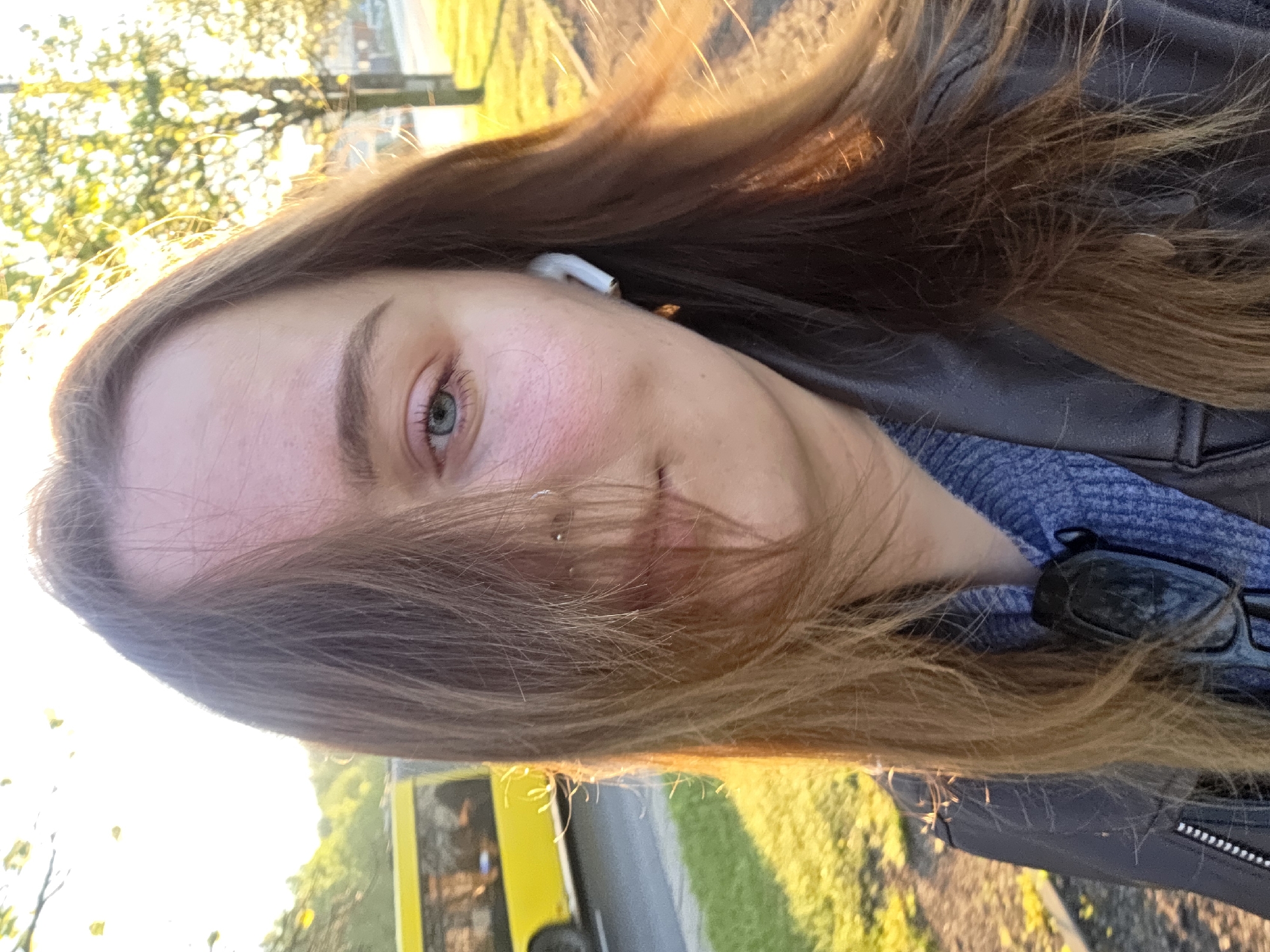 A close-up, portrait-style photograph depicts a person with long brown hair, wearing a white earbud in their left ear, and a blue shirt and black jacket. They appear to be outside, near a street with a yellow bus in the background. The person is looking at the camera, and their face is partially illuminated by sunlight. The photo seems to have been taken at eye-level, with a slight tilt. The specific location in the city of Unknown, Unknown is difficult to determine from the visual evidence.