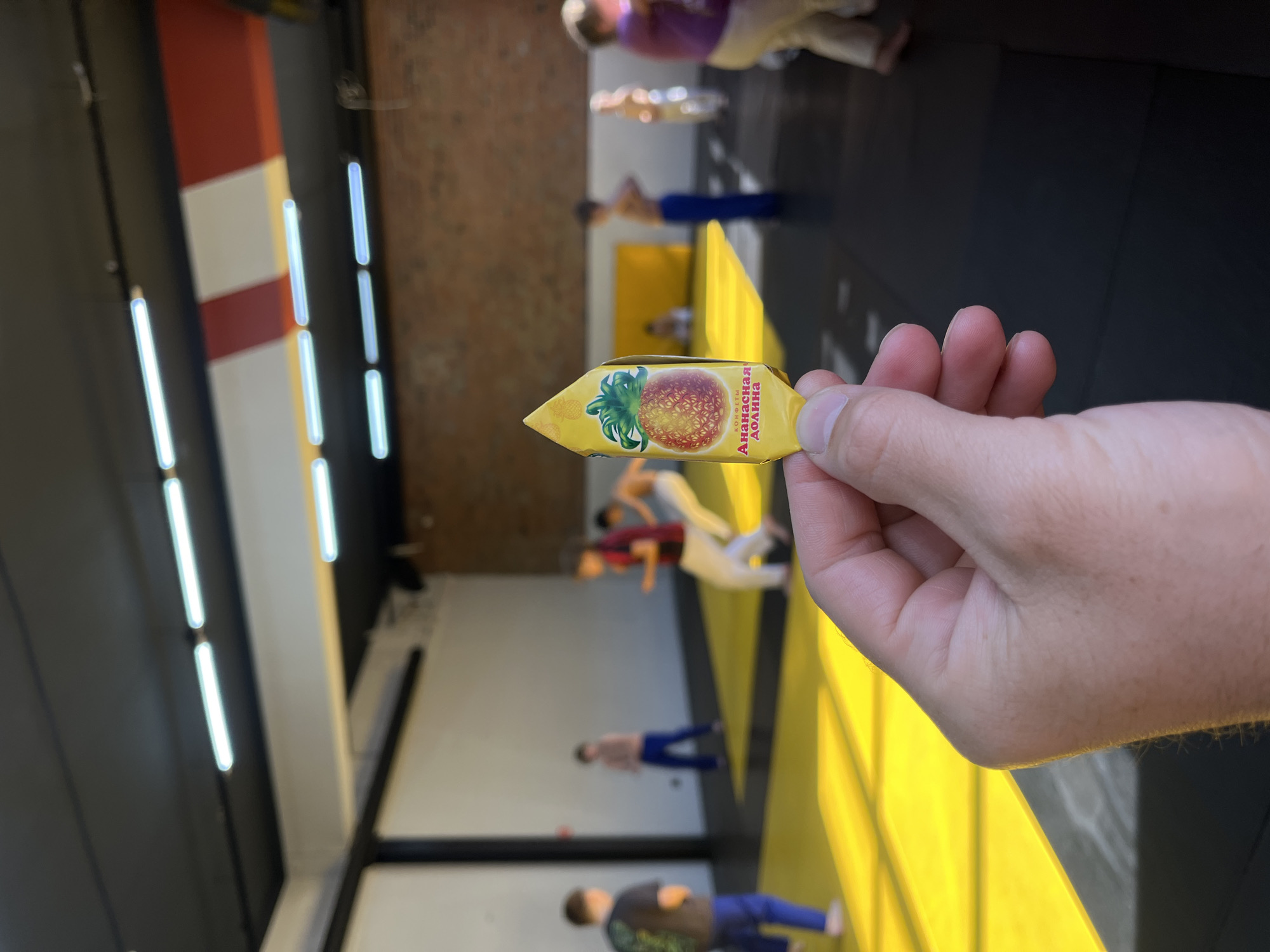 The image, taken in an indoor setting likely in Krasnoyarsk, Russia, captures a person's hand holding a candy wrapper in the foreground, with several blurred figures engaged in an activity in the background.
In the foreground, a right hand holds a yellow, arrow-shaped candy wrapper. The wrapper features a clear image of a ripe red strawberry. Visible text on the wrapper, written in Cyrillic, reads "КОНФЕТЫ" (Candies) and "ЗЕМЛЯНИЧНАЯ ДОЛИНА" (Strawberry Valley).
The background reveals a large indoor space, possibly a gym or a training hall. The floor is partially covered with a bright yellow mat, which transitions into a darker floor surface. Several out-of-focus individuals, appearing to be adults or older children, are spread across the yellow mat. They are dressed in various colored athletic wear, including purple, white, red, blue, and black, and appear to be standing in different stances, suggesting participation in a group activity, such as martial arts or exercise. The walls of the facility are dark grey or black, with some sections featuring distinctive red and white vertical stripes. Bright, white vertical strip lights are mounted on the walls, providing illumination to the interior space. The overall impression is one of an active training session.