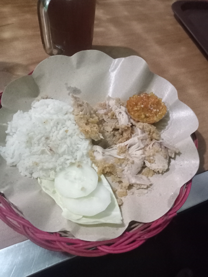 A prepared meal is depicted from an overhead perspective, presented on a light brown, scalloped-edge paper liner, which rests inside a woven red plastic basket. The scene is located in Ganung Kidul, Indonesia. The meal consists of a mound of white steamed rice positioned on the left side of the paper. Adjacent to the rice are pieces of shredded or pulled chicken, exhibiting both plain and lightly breaded textures. Above the chicken, a spherical mass of orange-yellow, textured paste, consistent with *sambal*, is present. Below the rice, two round, thin slices of white cucumber and a single light green cabbage leaf are visible. In the upper left background, a blurred clear glass containing a dark brown liquid is present. A dark brown surface is partially visible in the upper right background. The meal is situated on a wooden tabletop, with a metallic surface visible in the lower left foreground. No individuals are present, and no active interactions are depicted.