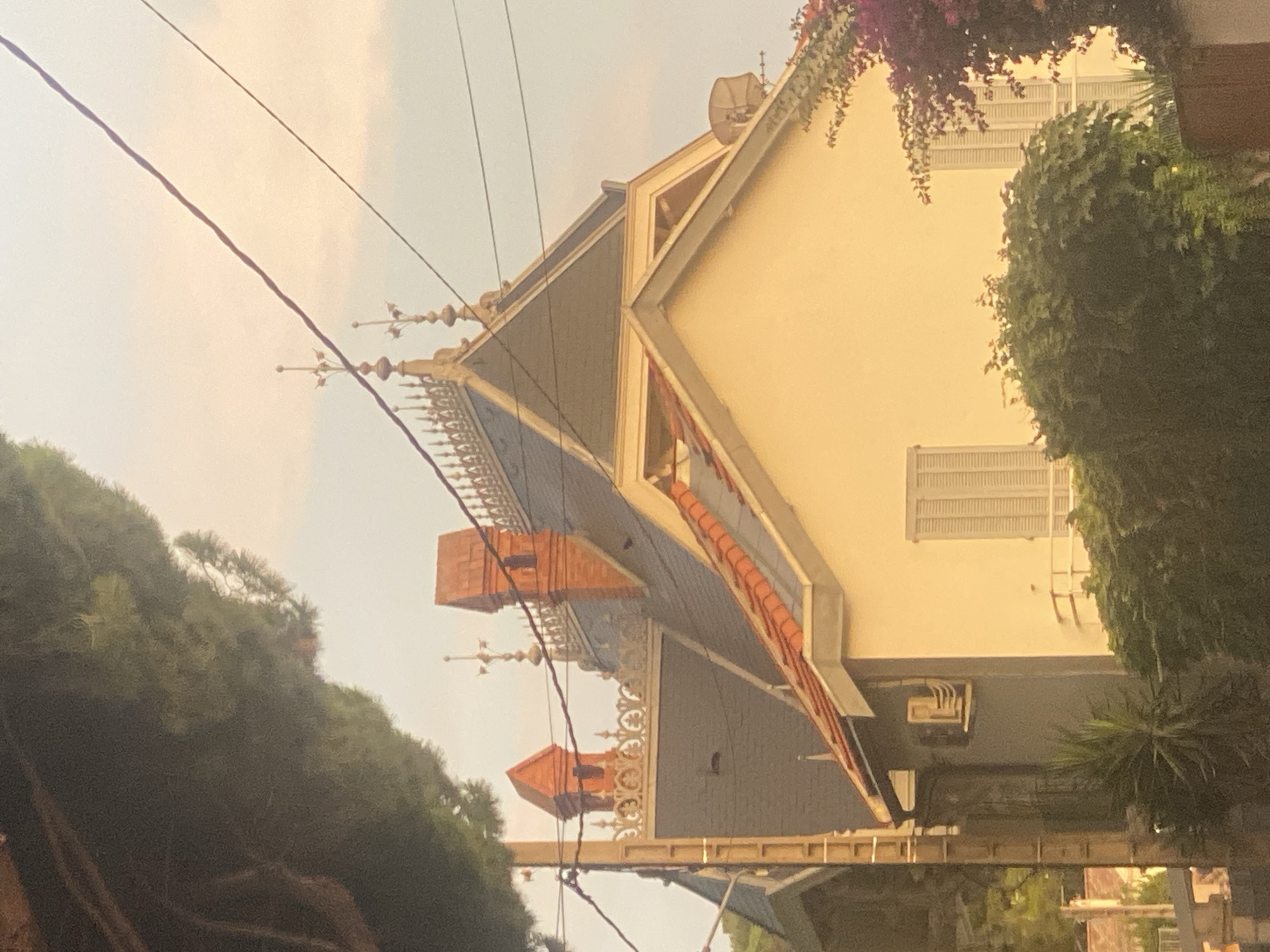 This is a photograph of a house located in Antibes, France, viewed from a low angle. The house has a light yellow facade with white shutters on the windows and is adorned with a red-tiled roof and ornate architectural details. The roofline features several chimneys and decorative ironwork. Lush green foliage, including bushes and flowering vines, surrounds the base of the house. A tall evergreen tree occupies the left side of the frame, partially obscuring the view. Overhead, power lines crisscross the sky, which is a soft pastel shade of orange and blue, indicating either sunrise or sunset. No people are visible in the image. The scene appears to be a residential area, characterized by its architecture, vegetation, and the presence of urban infrastructure like power lines.