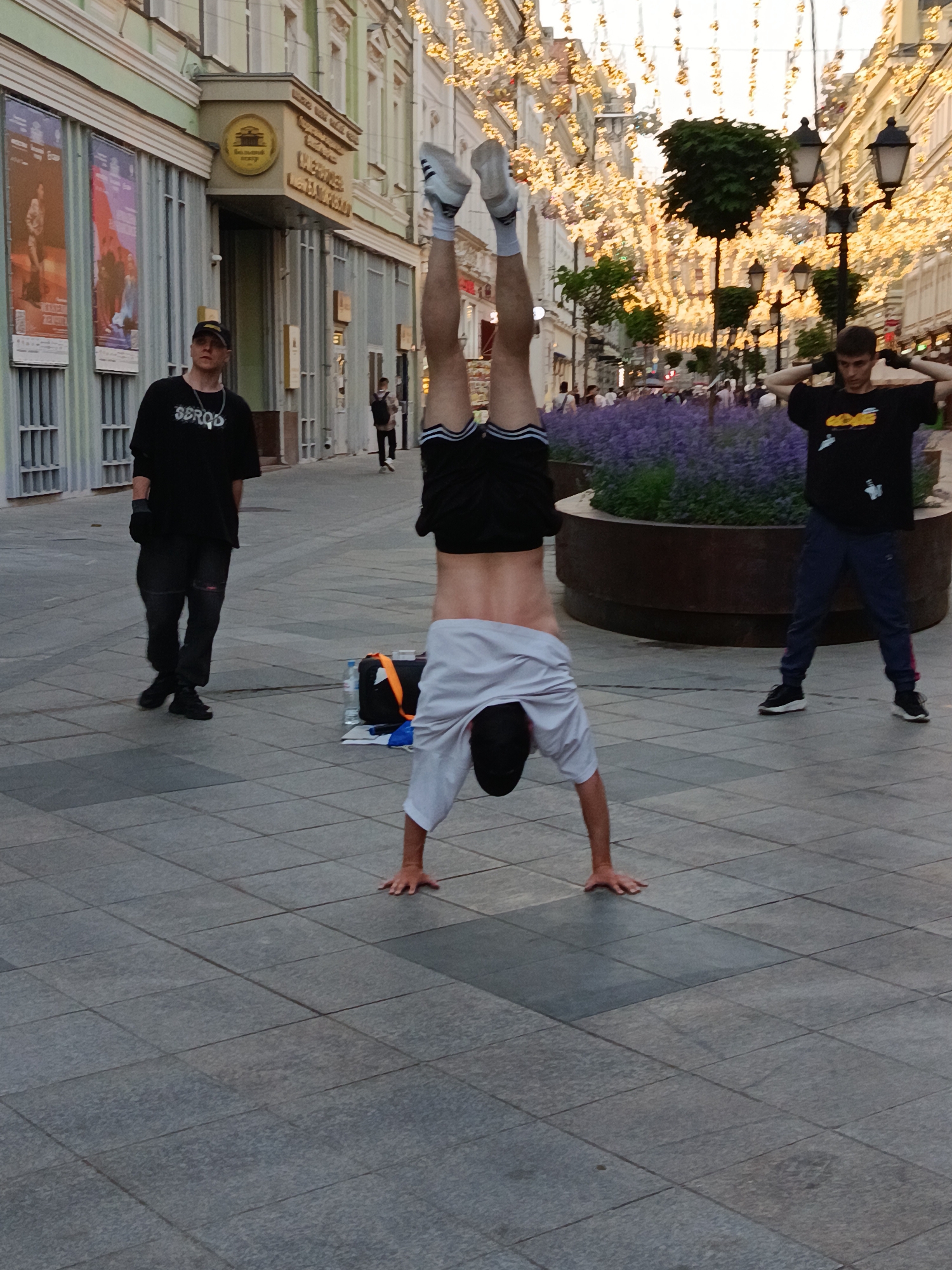 An individual is performing a handstand on a tiled pedestrian street in an urban environment, with their feet in the air and body inverted. The person is wearing a white t-shirt and black shorts with white stripes. Another person in a black t-shirt and dark pants stands to the left, wearing a black baseball cap and a necklace. A third individual in a black t-shirt and dark blue pants is positioned further to the right, with their hands behind their head. In the background, buildings with ornate facades are visible, decorated with numerous strings of golden lights. A planter filled with purple flowers and several lampposts are also present. A bag and a water bottle are on the ground near the handstand performer.