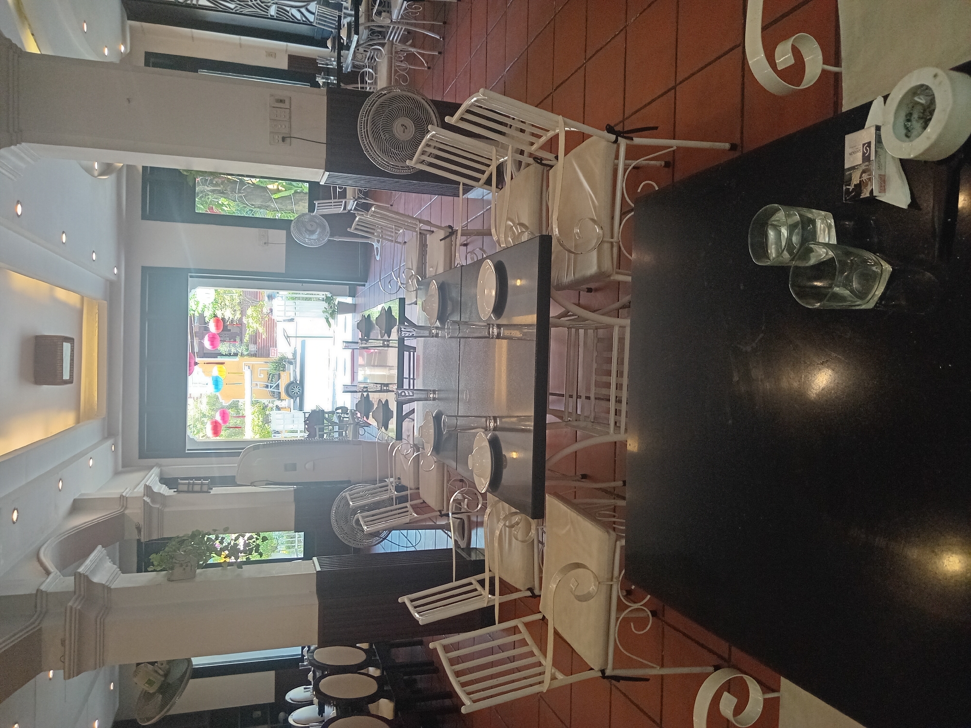 The image depicts the interior of a restaurant in Hoi An, Vietnam, with a focus on a dining area. White metal chairs with beige cushions are arranged around dark rectangular tables. Plates and glasses are set on some of the tables, indicating readiness for dining or recent use. The floor is covered with reddish-brown tiles. In the background, an open doorway reveals a sunny outdoor area with trees, colorful lanterns hanging, and a glimpse of vehicles. Two large circular fans are visible in the restaurant, one near the entrance and another positioned to the right. The architecture features prominent white columns and a decorated ceiling with recessed lighting. In the foreground, a close-up view of a dark table shows two glasses of water, a pack of cigarettes, and an ashtray with cigarette butts. The overall scene suggests a casual dining establishment in a warm climate.