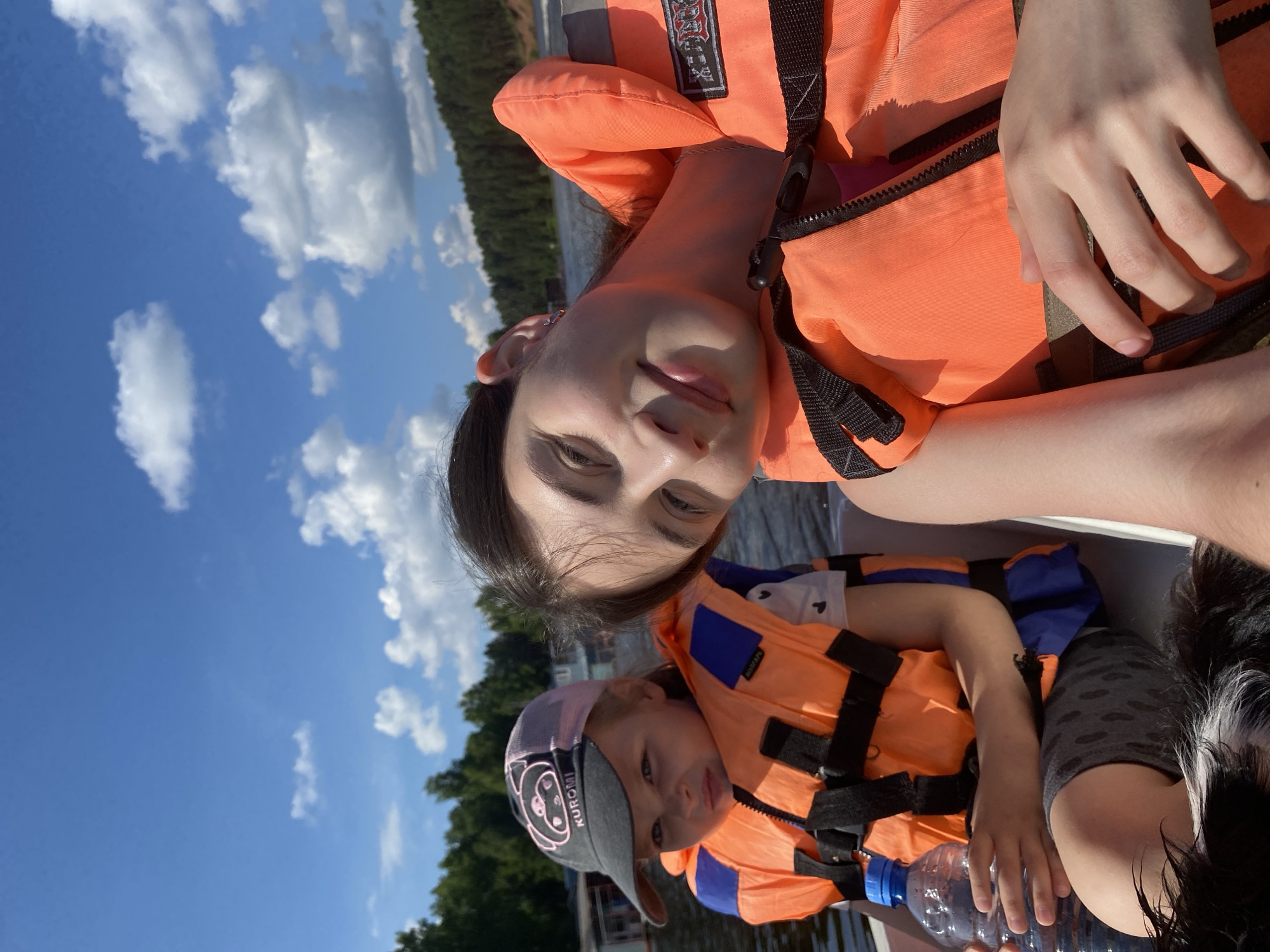 In the city of Kamakoye, Russia, two young females are captured in a close-up, selfie-style shot while on a boat. The girl in the foreground, on the right, is leaning back with a relaxed smile, her head resting on her shoulder. She wears an orange life vest over a pink garment. The girl on the left, younger in appearance, is looking towards the camera with a neutral expression, also wearing an orange life vest over a white shirt. She has a black baseball cap with a pink brim and the word "KUROMI" embroidered on it. A blue plastic water bottle is visible in front of her. Both individuals are seated on a white boat, with the background showing a body of water, a tree-lined shore, and a bright blue sky with fluffy white clouds. The image appears to have been taken on a sunny day.
