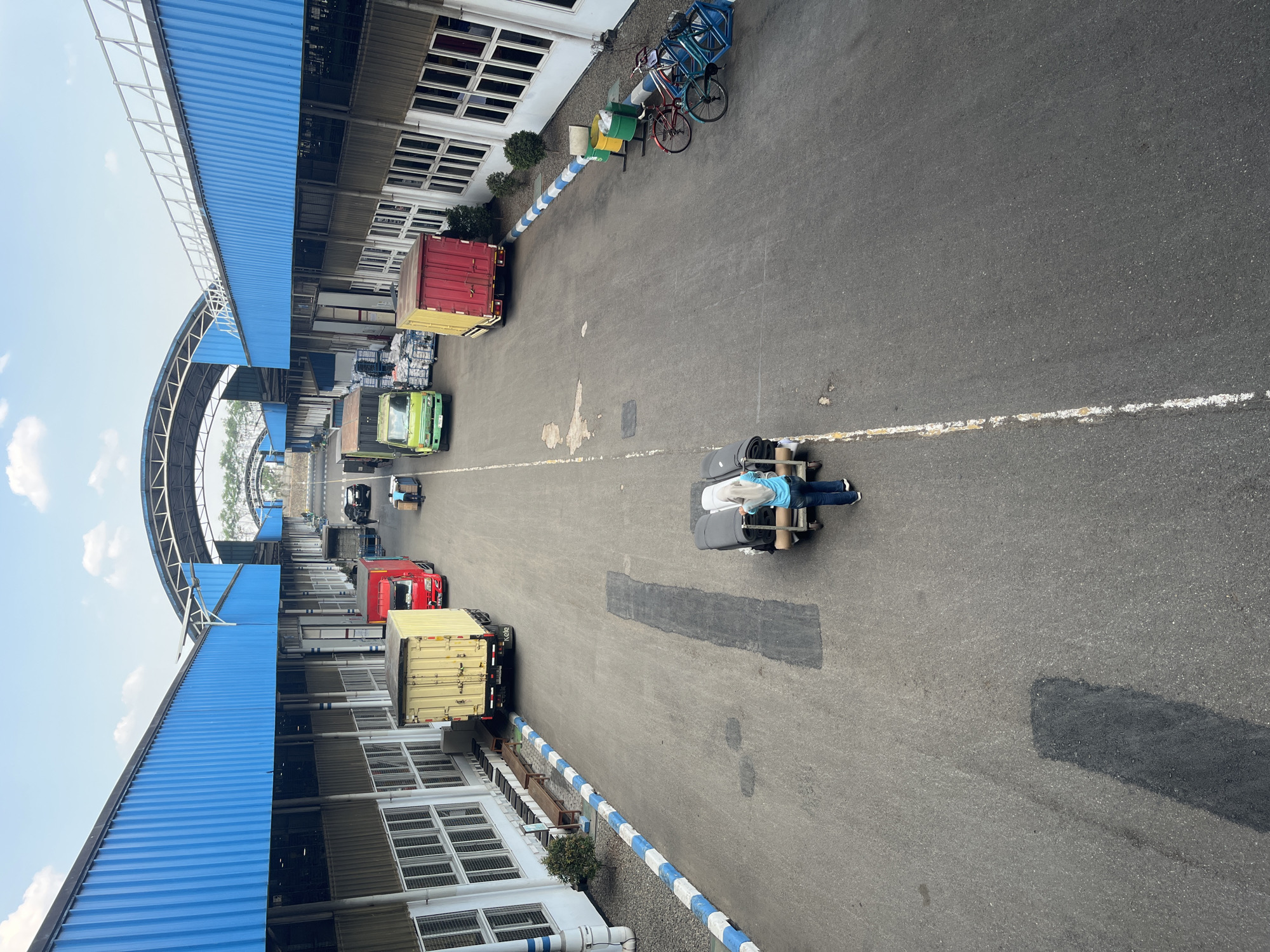 The image depicts an outdoor loading dock area in Pelang, Indonesia, likely at a warehouse or distribution center. In the foreground, a woman wearing a blue hijab and jeans is pushing a trolley loaded with rolled-up items, possibly fabric or carpets. She is on an asphalt road marked with a white line.
Several trucks are parked or moving in the background. On the left, a yellow box truck is parked. To its right, a red truck is visible, followed by a green box truck. Further down the road, another yellow box truck is parked, and a red truck is seen. Two cars are also present further in the distance.
The loading dock area is flanked by buildings with blue corrugated metal roofs and tan walls with rows of windows. There are white and blue striped curbs along the sides of the road. In the right background, two bicycles are parked near some green trash bins. The sky above is blue with scattered clouds. The overall scene suggests a logistics or transportation hub during daylight hours.