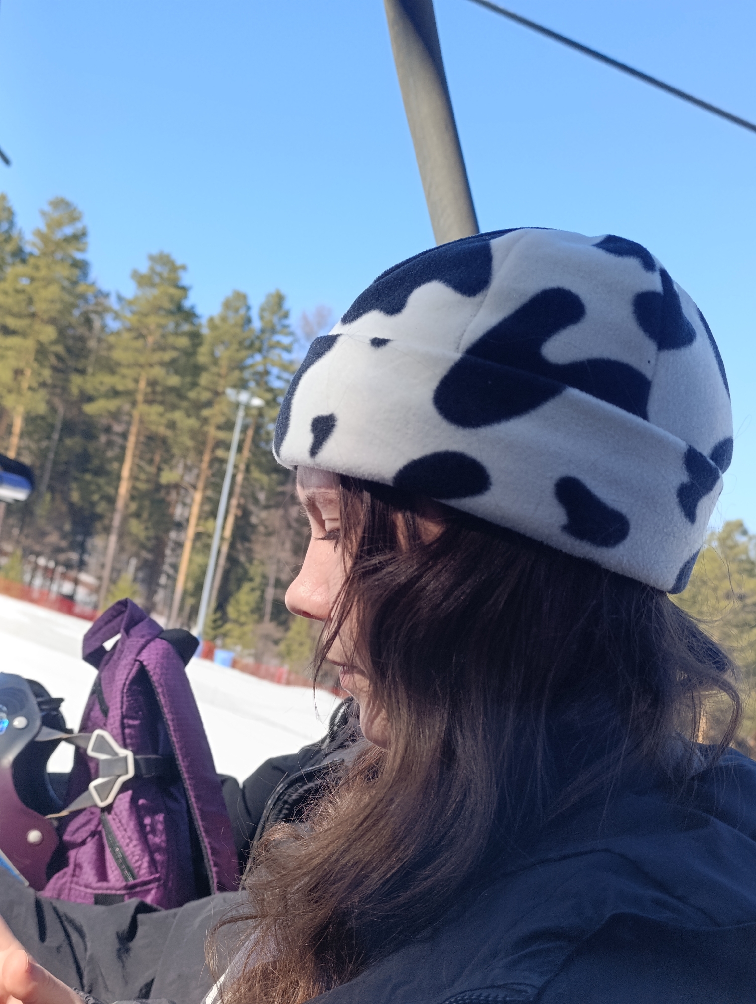 A close-up, daytime shot focuses on a person with dark hair, wearing a white hat with black cow spots, in what appears to be a winter outdoor setting. The person is positioned to the left, viewed from the side, with their face in profile. The subject's nose is prominent, and they are looking downwards. The individual is wearing a dark jacket, and a purple backpack is partly visible to their left.
The background features snow-covered ground and trees, suggesting a snowy or winter environment. A ski lift support is visible behind the subject and to the right. The sky is blue.
Based on the landscape and equipment present (ski lift), the location appears to be a ski resort in or near Krasnoyarsk, Russia. The scene captures a moment of outdoor activity, possibly involving winter sports.