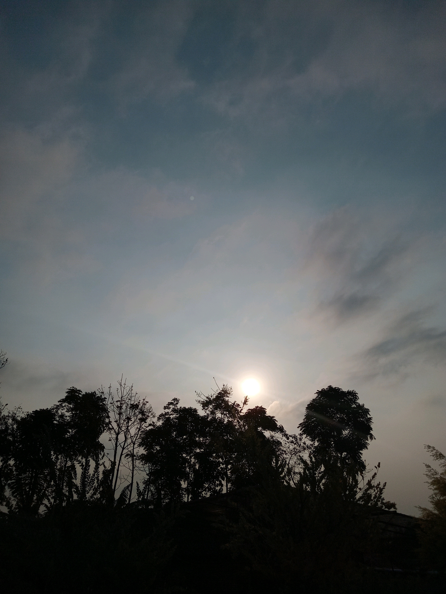 The image shows a silhouette of trees against a sky with a bright sun. The sun is partially obscured by clouds, casting a hazy glow. The trees are dark and appear to be dense, with some bare branches visible among the foliage. The sky is a gradient of blues and grays, with wispy clouds that suggest it may be dawn or dusk. The overall impression is one of natural beauty and tranquility. The image was taken in Claket, Indonesia.