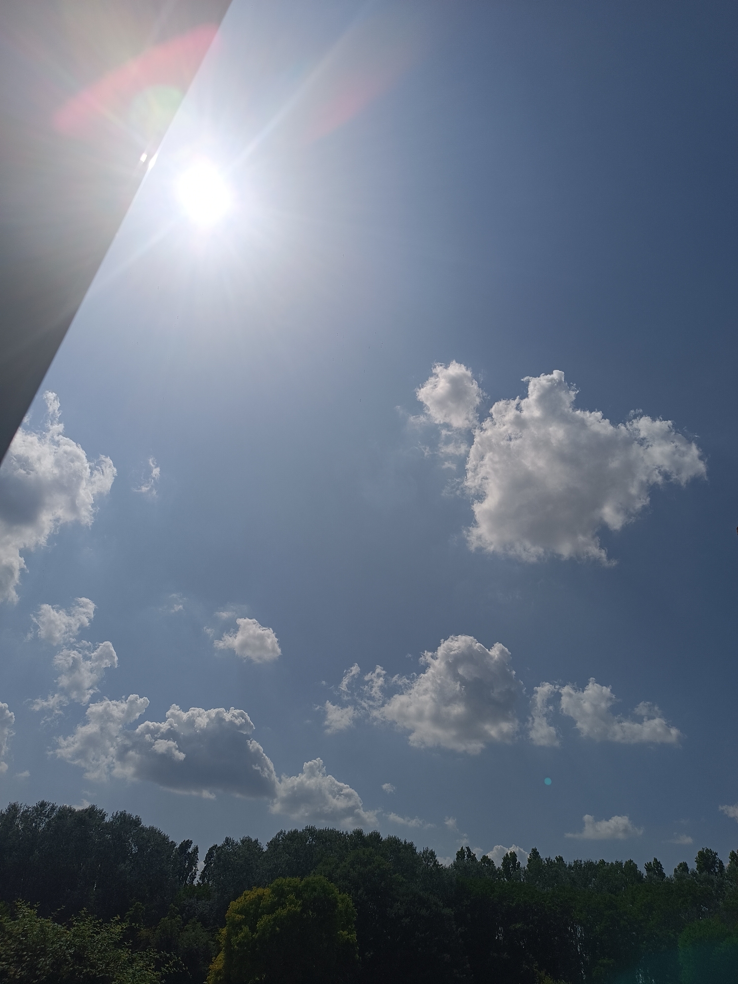 The image shows a bright, sunny sky with scattered white clouds. The sun is a prominent feature, shining brightly in the upper left portion of the frame, causing lens flare with visible streaks of light and colorful reflections. A dark, angled object, possibly a building or structure, partially obstructs the view of the sun from the left side. Below the sky, a dense line of trees forms a dark green horizon, suggesting a wooded area or park. The overall scene is outdoors, under a clear blue sky with sunlight. There are no people or specific landmarks visible that would indicate a precise location within Casalmaggiore, Italy, beyond it being an outdoor scene with trees and sky.