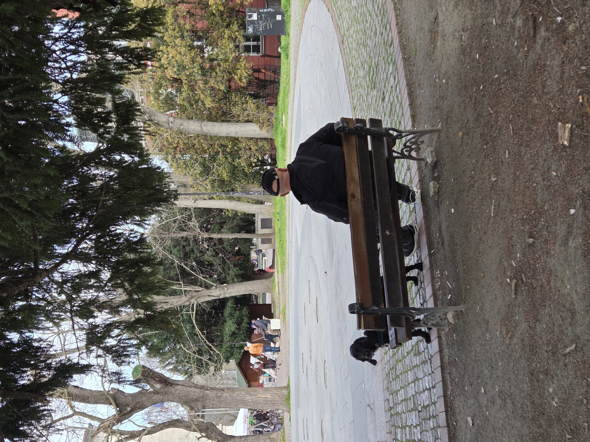 A man in a black jacket and cap sits on a wooden bench in a park. The bench is located on a paved pathway with a brick border. The ground beside the path is littered with debris and cigarette butts. In the background, several people are congregated around a small building or stand. Trees surround the area. The scene is likely in a public park or garden in Varna, Bulgaria.