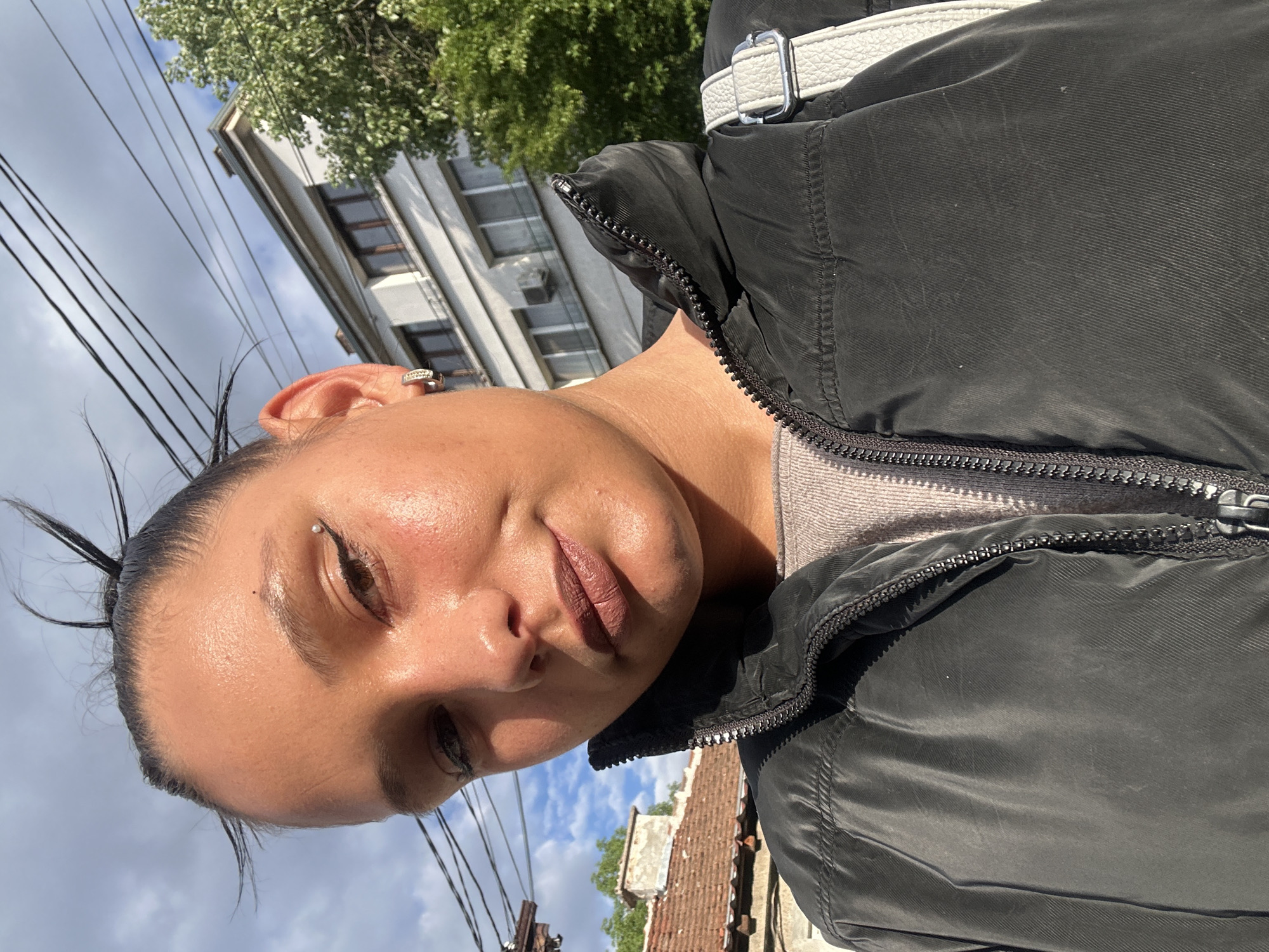 A selfie of a woman is taken outdoors. She has fair skin, brown hair pulled back, and a visible piercing above her left eye. She is wearing a black puffer jacket over a light grey shirt.
The background includes urban elements. Behind the woman is a multi-story building with visible windows and air conditioning units. Above and to the left of the building are power lines and a tree with green leaves. To the lower right, a glimpse of a rooftop with red tiles can be seen. The sky, visible in the background, has a mix of blue and clouds.
Based on the architecture, urban environment, and power lines, it's possible this photo was taken in a residential area of Sofia, Bulgaria.