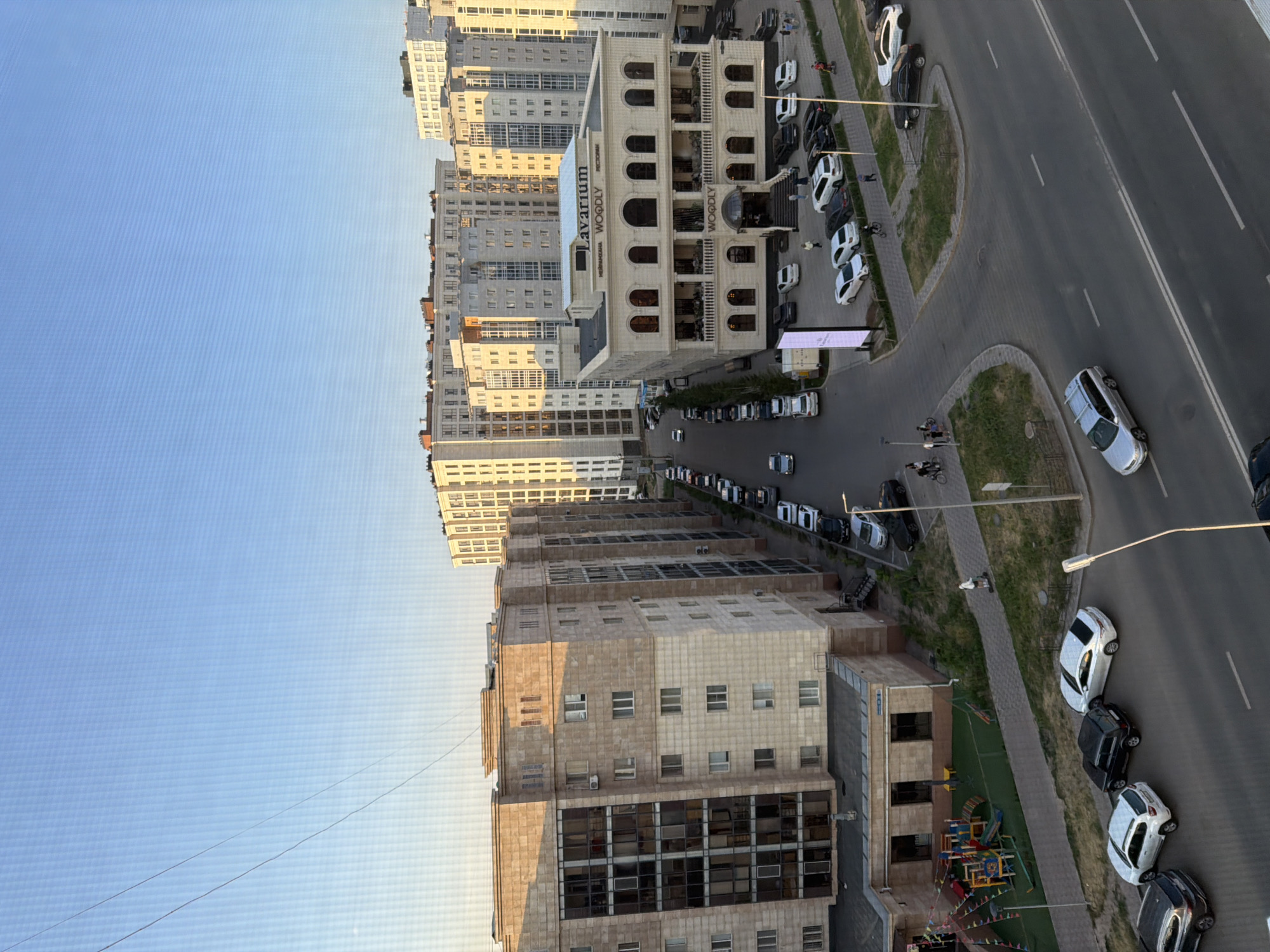 The image depicts an urban street scene in Astana, Kazakhstan, with modern residential buildings and a commercial establishment. In the foreground, a grey station wagon is driving on the right side of a two-lane road. Further down the road, several cars are parked along the curb, including white sedans, a black SUV, and a silver sedan. A playground with colorful equipment is visible on the left side of the street, adjacent to a tan-colored apartment building. Across the street, a multi-story building with arched windows and a sign that reads "Lavarium WOODLY" is prominent. This building appears to be a restaurant or entertainment venue. Behind it, numerous high-rise apartment buildings with light-colored facades and balconies line the horizon, characteristic of Astana's urban development. People are seen walking and cycling on sidewalks and near the buildings. The sky is clear and blue, indicating daytime. The overall impression is of a contemporary urban environment.