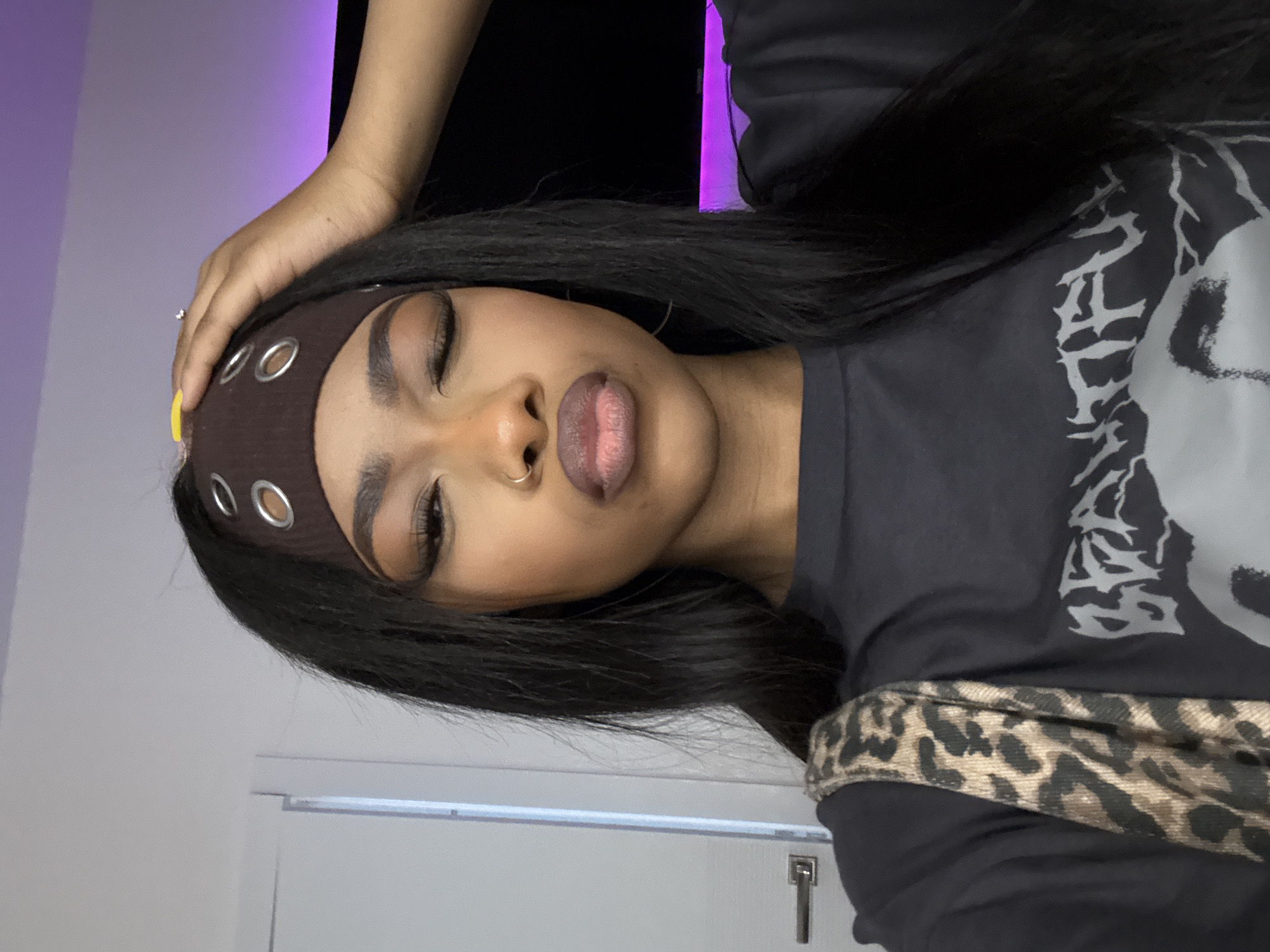A medium-shot of a person, likely female, with tan skin and long black hair wearing a brown headband with metal grommets. Her eyes are partially closed and she has a septum piercing. She wears a dark t-shirt with a light grey graphic and a leopard print strap is slung across her chest. The person's right hand touches her head. The background includes a white wall with a door and a gradient of black and purple. There is no explicit indication that this image was taken in Troitsk, Russia.