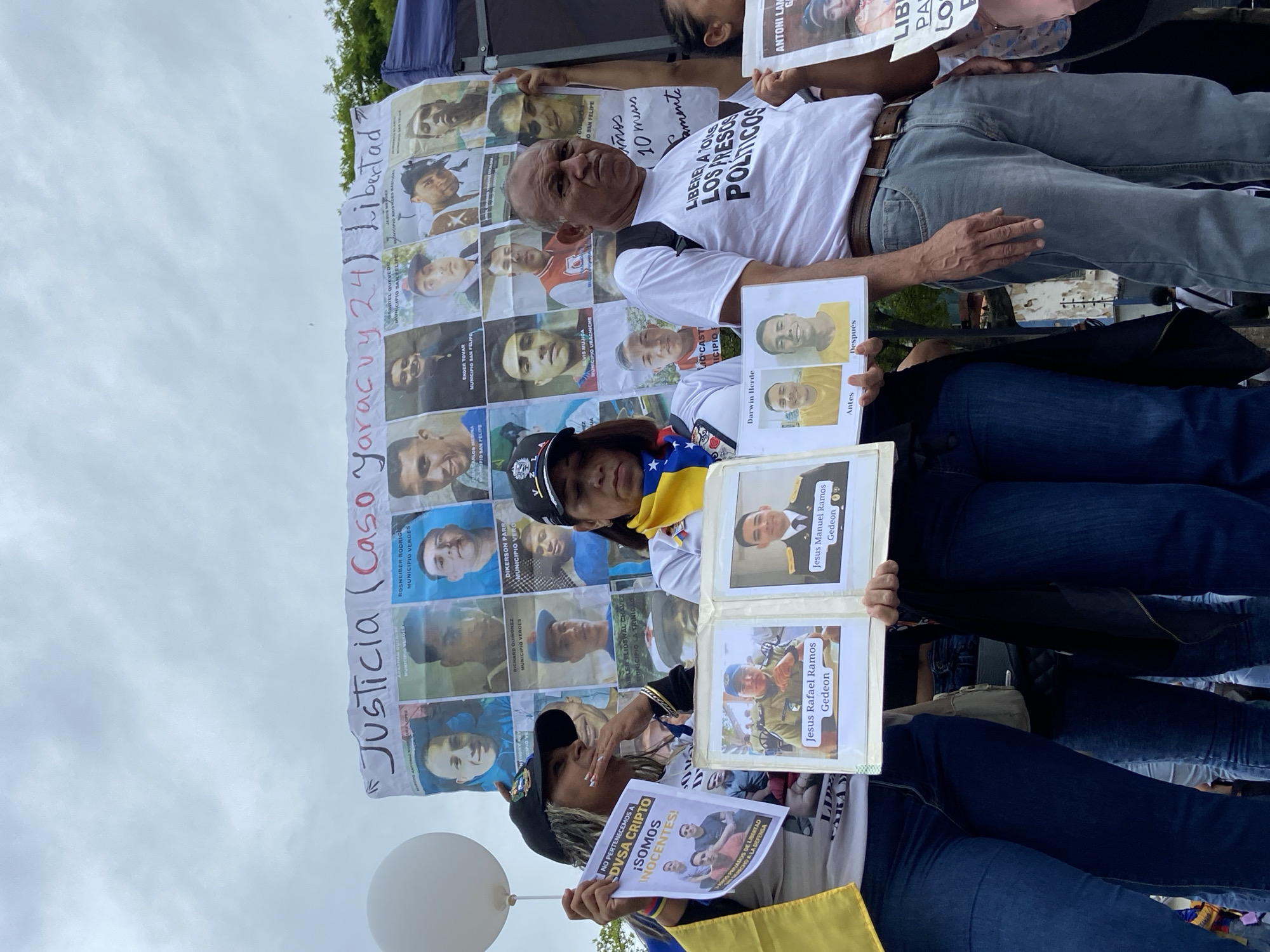 This image captures an outdoor demonstration in Caracas, Venezuela, under an overcast sky during the daytime. Several people are gathered, holding signs and banners, advocating for justice and the release of political prisoners.
In the foreground, a man in a white t-shirt stands prominently, holding a large white banner covered with numerous passport-style photos of men. Above these photos, red text reads "Justicia (Caso Yaracuy 24) Libertad" (Justice (Yaracuy Case 24) Freedom). Some names and municipalities like "ROSNEIBER RODRIGU MUNICIPIO VEROES" and "GABRIEL QUEVEDO MUNICIPIO SAN FELIPE" are visible alongside the faces. The man's t-shirt explicitly states "LIBERET A TOLLS LOS PRESOS POLITICOS" (Liberate all political prisoners).
To the right, a woman wearing a black cap and a white t-shirt, with a Venezuelan flag draped around her neck, holds an open album. Inside are photos, including "Antes" (Before) and "Después" (After) images of "Darwin Herde," as well as pictures of "Jesus Rafael Ramos Gedeon" and "Jesus Manuel Ramos Gedeon." Another woman, partially visible to the left, holds a white balloon and a small sign that reads "NO PERTENECEMOS A DVSA CRIPTO ¡SOMOS INOCENTES!" (We do not belong to DVSA CRIPTO We are innocent!), along with "NGO PRIVADOS DE LIBERTAD" (NGO Deprived of Liberty) and "CHO A LA DEFENSA" (Right to Defense). Another sign partially visible mentions "ANTONI LAM." The overall atmosphere conveys a somber but determined demand for human rights and judicial fairness.
