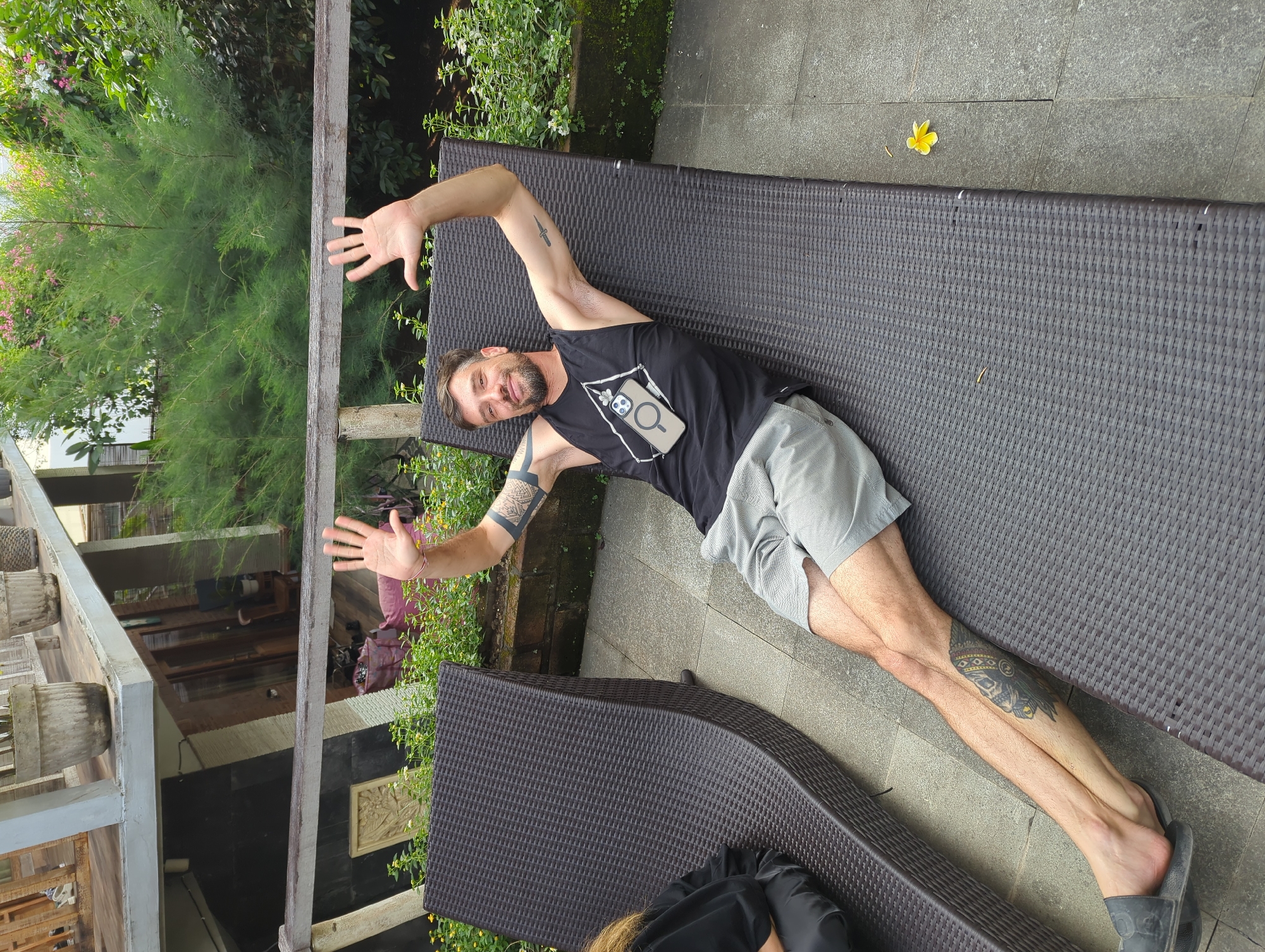 A man is lying on a dark brown wicker-textured chaise lounge on a stone patio in North Kuta, Indonesia. He is wearing a black tank top with a white graphic design, gray shorts, and a dark gray slipper on his right foot. The man has fair skin, dark hair, a beard, and tattoos on both arms and one on his lower right leg. He has his arms raised and hands open as if he is gesturing. The chaise lounge is partially shaded. A fallen yellow flower lies on the patio beside the lounge. In the background are lush green plants and a building with a balcony.