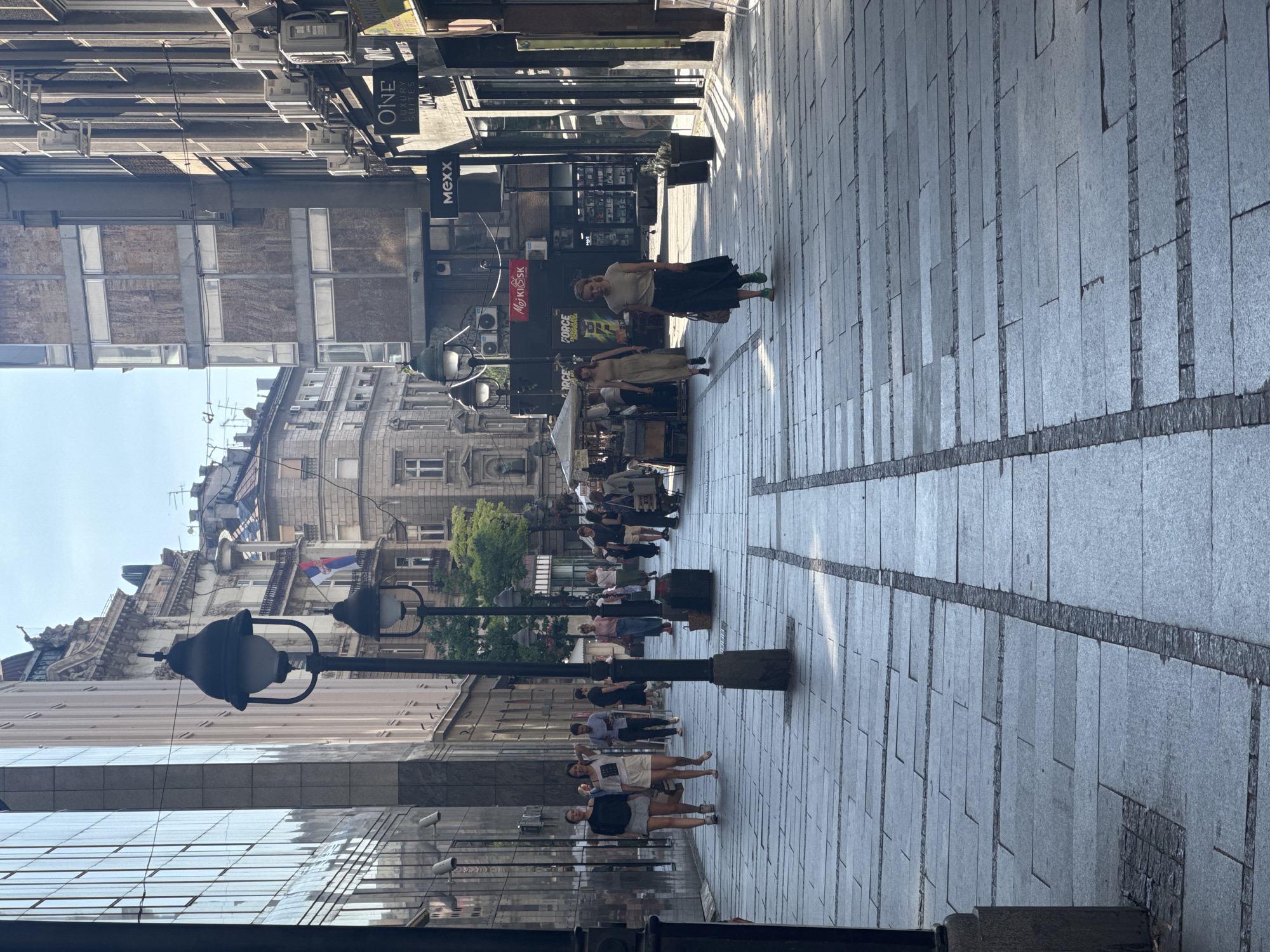 The image depicts a sunny day on a wide pedestrian street in Belgrade, Serbia, likely Knez Mihailova Street, known for its shops and vibrant atmosphere. The street is paved with large gray stone tiles, laid in a pattern with narrower dark stone borders. Several lampposts with a vintage design punctuate the scene. On the left, a modern building with a glass facade reflects the sky and surrounding architecture. Across the street, older European-style buildings with intricate details stand adjacent to more modern structures, some with visible air conditioning units.
In the mid-ground, several individuals are walking along the street. Two young women in casual attire are walking towards the viewer on the left side. Further down, a man in a blue shirt and dark pants is walking away. On the right side of the street, a woman in a dark skirt and light-colored top is walking towards the viewer, carrying a bag. Behind her, more people are seen strolling, some in groups. Various shop fronts are visible along the street, with signage including "ONE LUXURY SUITES" and "Mexx." A food stall with a canopy is also present. The overall impression is of a bustling urban environment with a mix of historical and contemporary architectural styles.