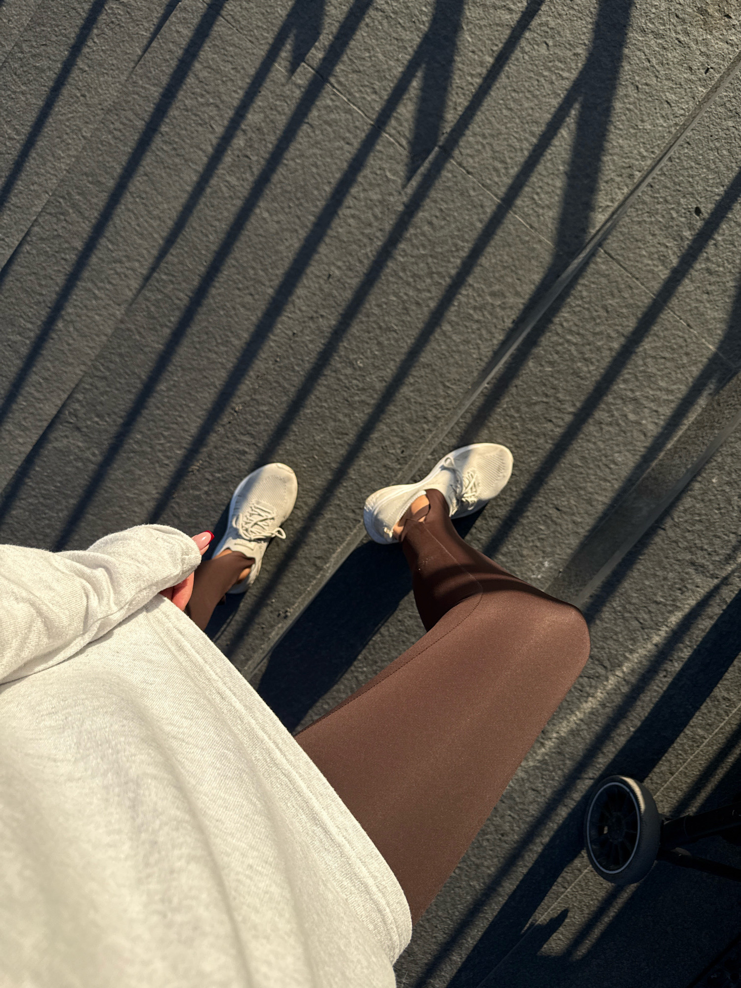A first-person perspective shot captures a person's legs and feet, dressed in brown leggings and white sneakers, standing on dark, textured steps. The steps are illuminated by sunlight, casting long, distinct shadow lines across the surface. A portion of a white, textured garment, likely a sweatshirt or jacket, is visible in the lower left corner. In the lower right, a black stroller wheel is partially visible. The scene appears to be outdoors, possibly in an urban setting with architectural elements like stairs. The image quality is clear, with good lighting and focus on the subjects. No specific location in Pavlodar, Kazakhstan can be determined from the visual information provided.