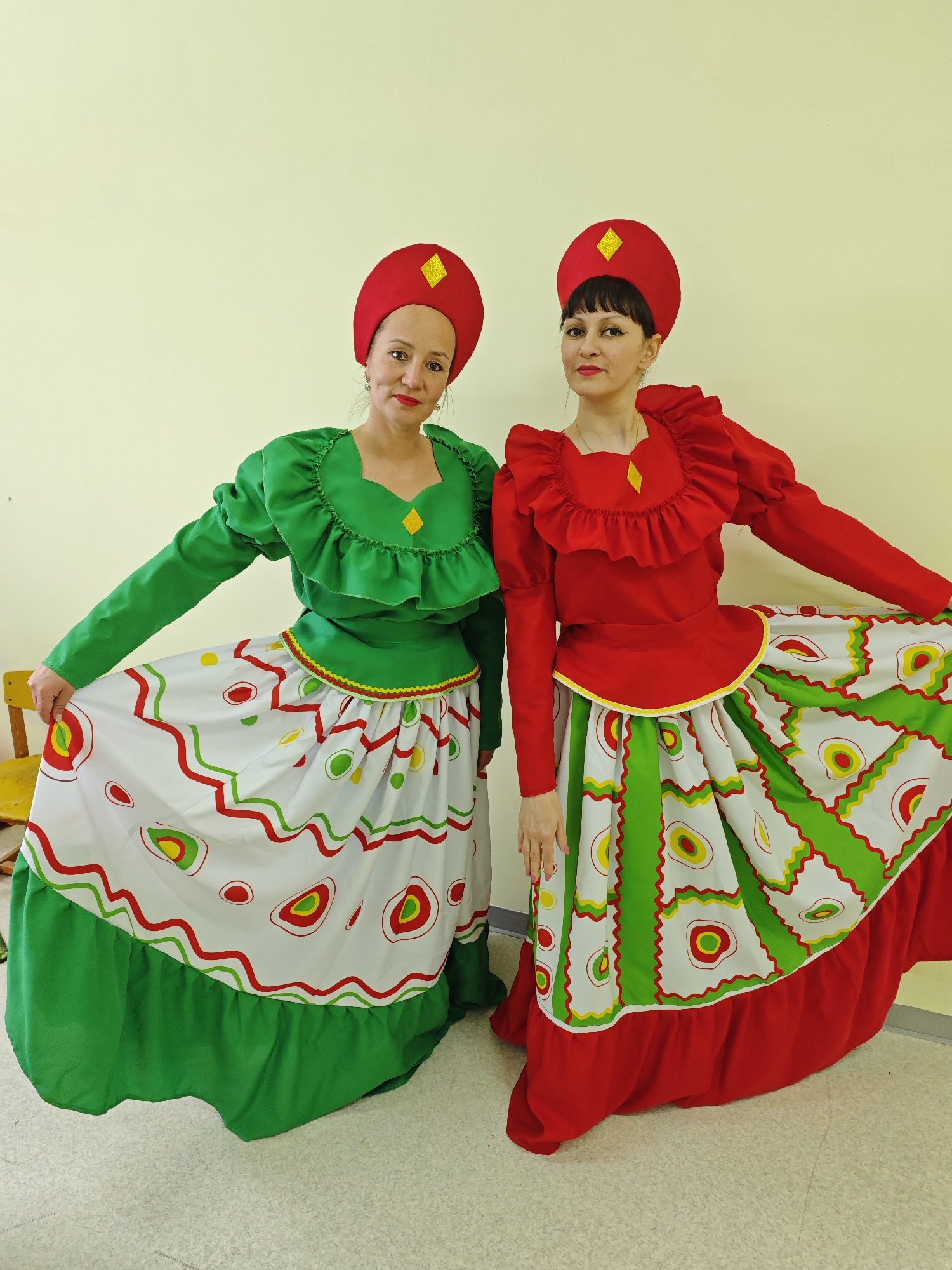Two women in brightly colored traditional-style costumes pose indoors against a plain light yellow wall in Markovskii, Russia. The setting appears to be a public or institutional building, possibly a community center or school, with a light-colored, patterned floor.
The woman on the left wears a vibrant green long-sleeved blouse with a ruffled neckline and a full white skirt featuring wavy green and red lines, and circular yellow, red, and green patterns. The skirt has a solid green ruffled hem. She completes her outfit with a red hat adorned with a yellow diamond-shaped embellishment. She looks towards the camera with a slight smile, holding up a portion of her skirt with her right hand. A small, yellow wooden chair is partially visible behind her.
The woman on the right wears a similar style, with a bright red long-sleeved blouse featuring more prominent ruffles around the neckline and shoulder area, and a full white skirt with wavy red and green lines, and circular yellow, red, and green patterns. Her skirt has a solid red ruffled hem. Like her companion, she also wears a red hat with a yellow diamond accent. She stands poised, looking at the camera with a neutral expression, holding up her skirt with her left hand.
Both women's costumes are elaborate and festive, suggesting they are prepared for a cultural performance, event, or a photoshoot showcasing traditional dress. The lighting is even and bright, typical of indoor artificial light. No text is visible in the image.