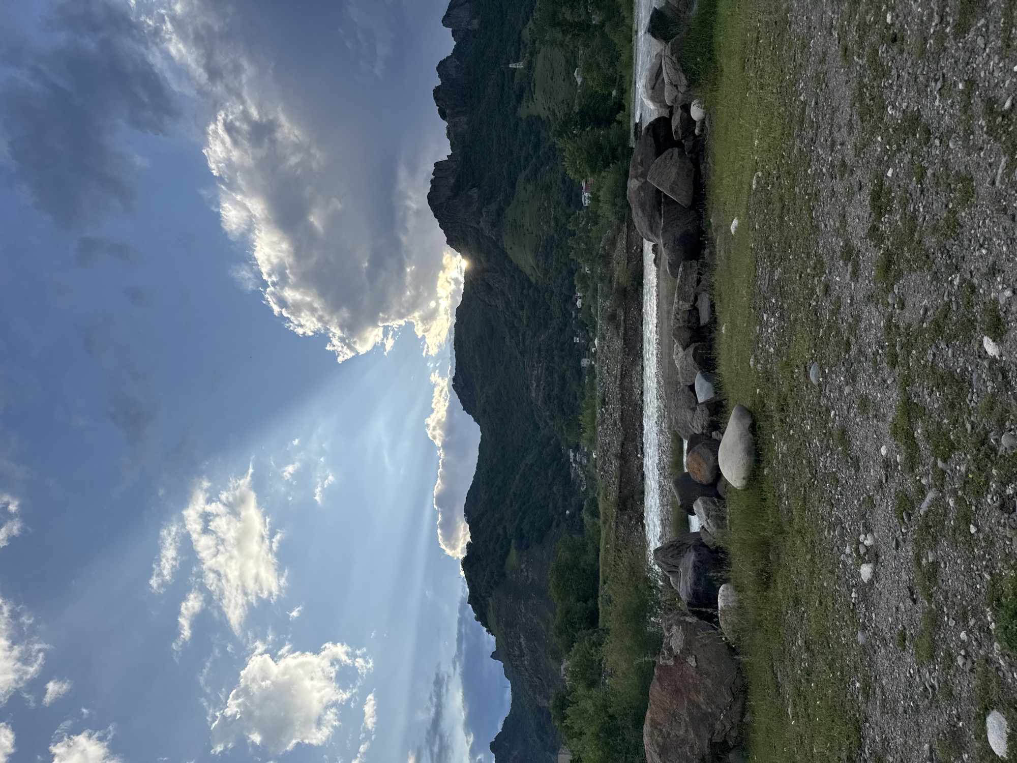The image depicts a scenic landscape featuring a river in the foreground, bordered by grassy terrain and large rocks. Beyond the river, a range of verdant, tree-covered mountains dominates the middle ground, with a small settlement visible nestled within the foothills. The sky above is a dynamic mix of blue sky and dramatic clouds, with sunbeams breaking through, suggesting late afternoon or early evening. The setting appears to be a natural, undeveloped area, likely a rural or mountainous region. The image quality is good, with clear details and natural lighting, consistent with a photograph taken outdoors. There are no people or animals visible in the frame. The location is described as being in the city of Karachayevsk, Russia, which is consistent with a mountainous terrain and possible settlement.