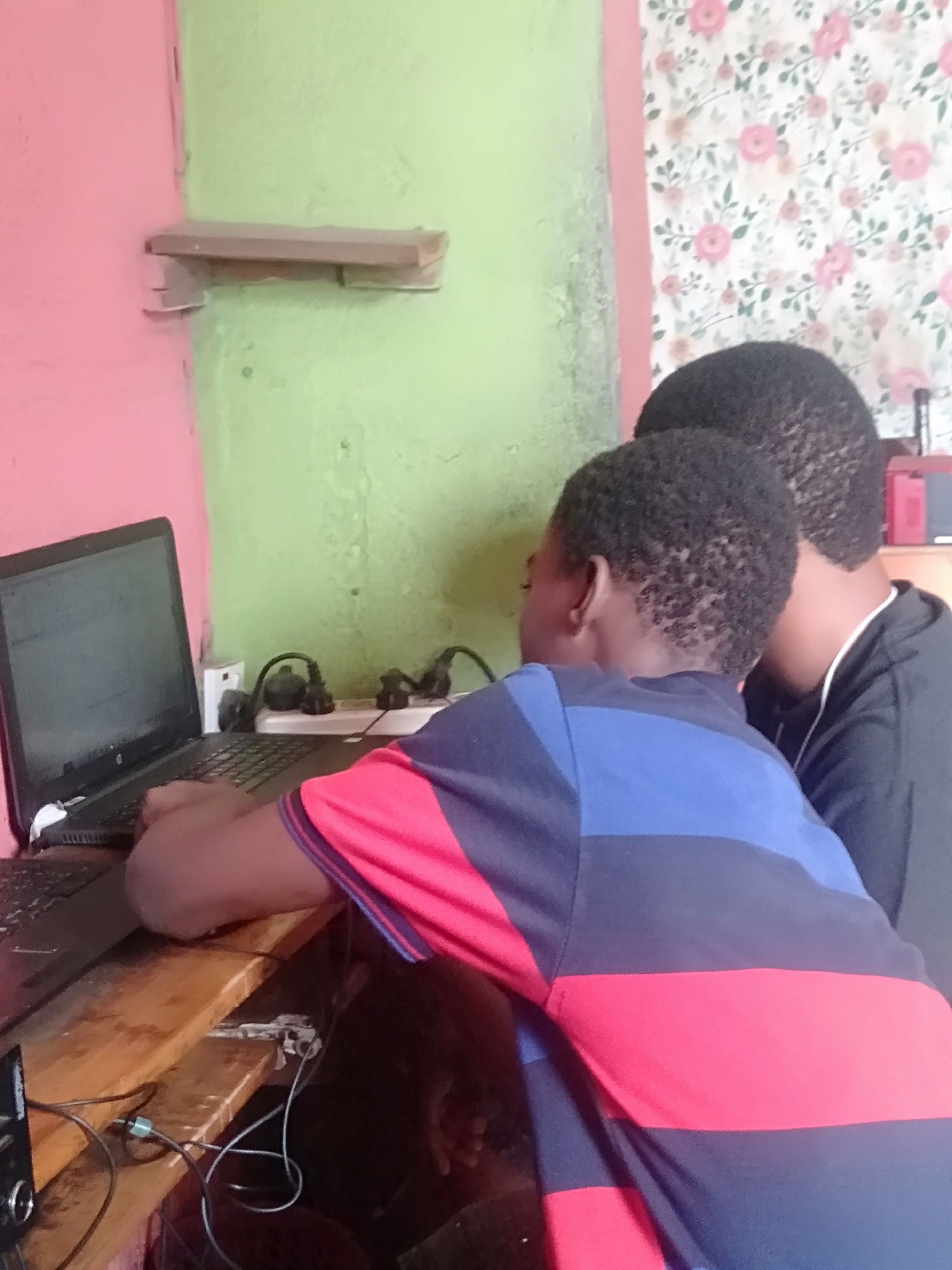 Two Black children are seated at a wooden desk using a laptop. An electrical outlet and a power strip are visible on the wall above the desk. A small, unpainted shelf is mounted on the wall. The walls are painted pink and green, with a floral wallpaper section. Cords from the electronics are tangled on the desk and floor. The location is identified as Shangisha, Nigeria.
