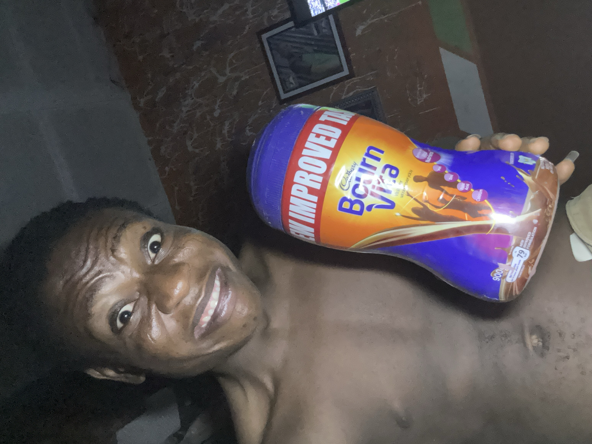 The image is a selfie taken by a man with dark skin, holding a large container of Cadbury Bournvita in his right hand. The container is mostly orange and blue, with text advertising it as "NEW IMPROVED T..." and "Bourn Vita". The man is smiling and his face is slightly angled towards the camera. He is shirtless and appears to be in a dimly lit room. Behind him, there's a wall with a patterned covering and two framed pictures hanging on it. A television screen is visible at the top, though its contents are unclear. The location is Unknown, Unknown.