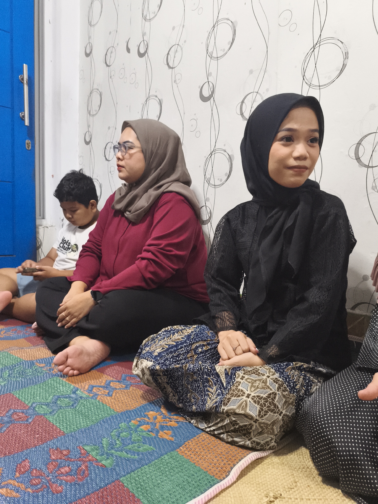 The image depicts an indoor scene in Depok, Indonesia, with four individuals seated on a colorful woven mat. In the foreground on the right, a woman wearing a black hijab and a black lace top is seated, looking to her left. To her left, another woman in a maroon long-sleeved top and a grey hijab is seated, also looking to her left. Behind them, a young boy in a white t-shirt with text is seated and looking down at a mobile phone. A fourth person, mostly obscured, is partially visible on the far right. The background features a white wall with a patterned wallpaper of black swirls. A blue door with silver handles is visible on the far left of the frame. The lighting appears to be artificial, likely from overhead sources.