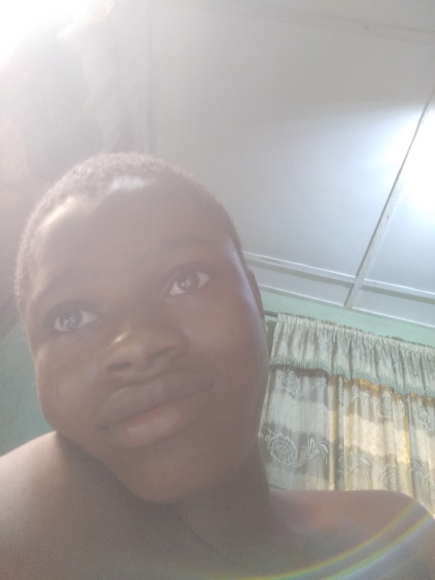 A close-up, high-angle photograph depicts a person with dark skin and short, dark hair. The individual is looking up and slightly to the left. Behind the person, there is a white ceiling with what appears to be a drop-down panel and a section of light-colored curtains with a fringed valance. A bright, diffuse light source is present in the upper left corner of the frame, and a rainbow-like effect is visible near the bottom of the image. The location is Unknown, Unknown.