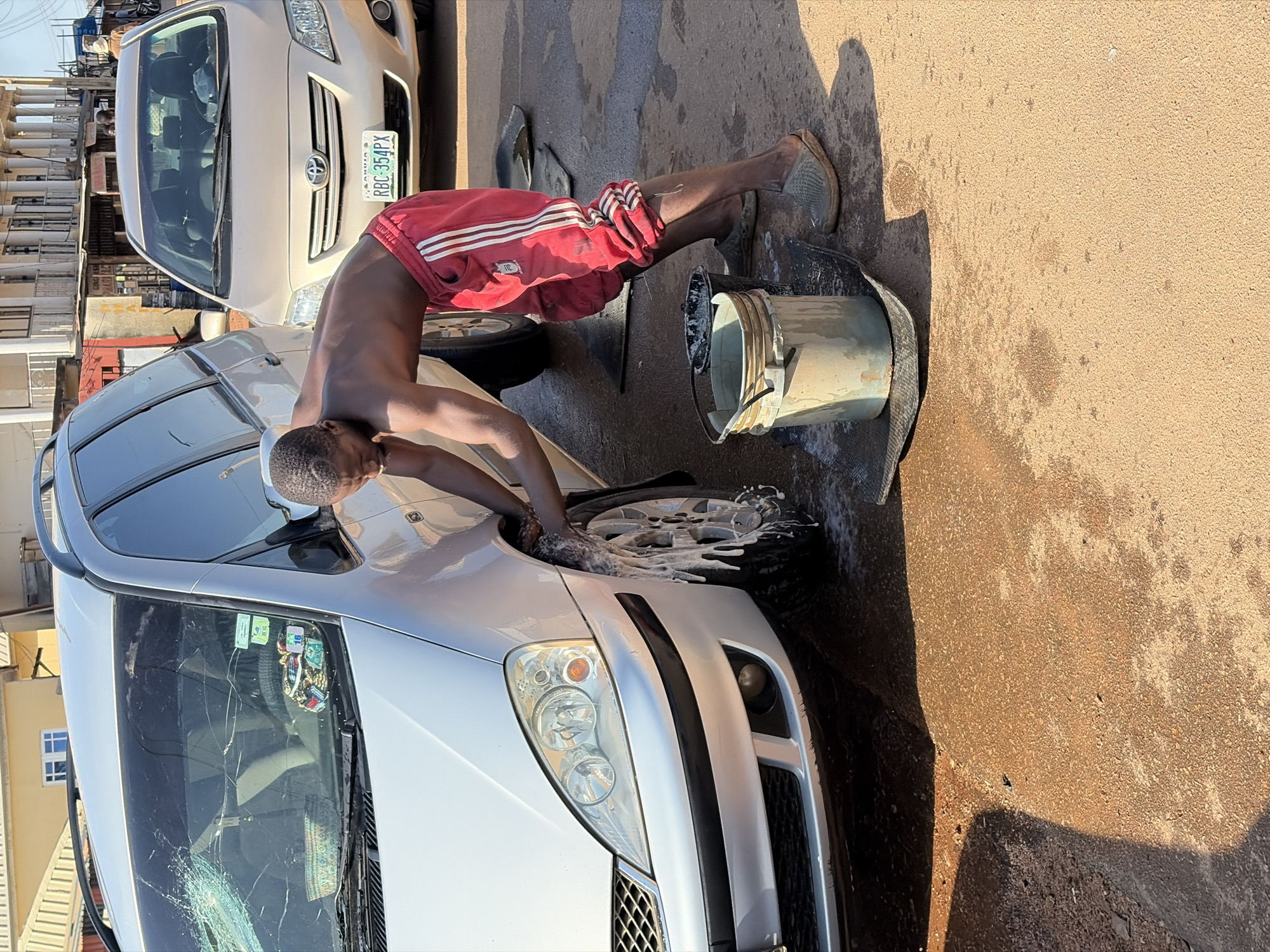A bare-chested young Black man, wearing red track pants with white stripes, is washing the front wheel of a silver minivan. He is using a sponge to scrub the tire, which is covered in white soap suds. Two buckets, one empty and one with soapy water, are on the ground beside him. A second car, a beige sedan with a license plate reading "RBC 354PX," is parked behind the minivan. The scene appears to be a street in Gura Top, Nigeria, with buildings visible in the background. The ground is wet and sandy, indicating the car wash is taking place outdoors. The minivan's windshield is cracked.