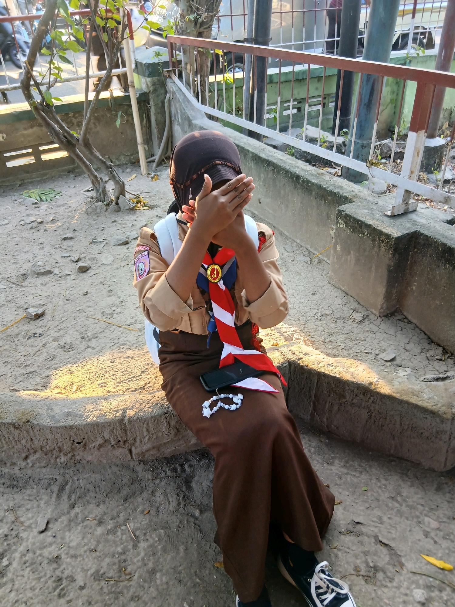 A person is sitting on a concrete ledge, covering their face with their hands. They are wearing a brown uniform with a sash and a headscarf, suggesting they might be a scout. A mobile phone and a white beaded bracelet are placed on their lap. The background features a rusty metal fence and some foliage, indicating an outdoor urban setting. The lighting suggests it's either early morning or late afternoon. The specific location in Gorontalo, Indonesia, cannot be determined from the image alone.