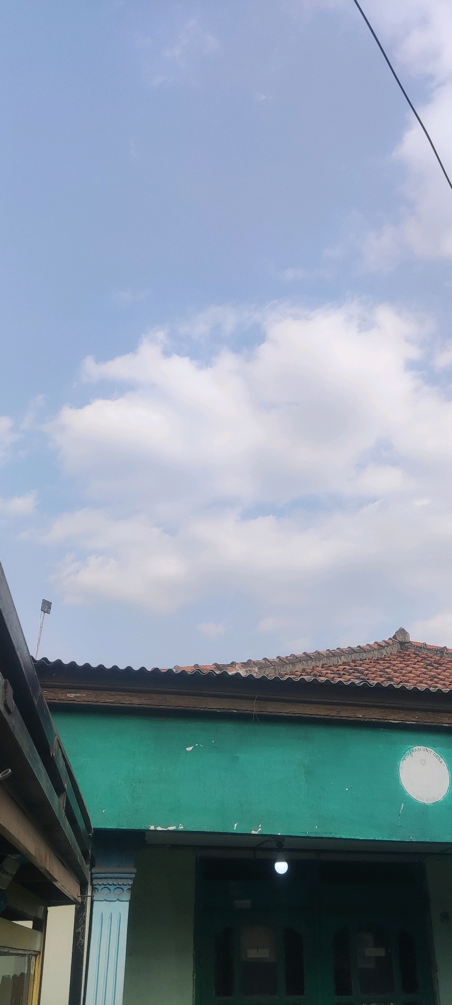 The image is an outdoor, upward-facing shot of a house and the sky. The sky is a light blue with scattered white clouds. A power line is visible diagonally in the upper right corner. The roof of a house, made of red tiles, is in the middle ground. The walls of the house are painted a vibrant green. There is a circular emblem on the green wall, to the right of the center, with text that is difficult to read. Part of a wooden structure is visible on the left side of the image. A small antenna is mounted on the roof. The lower part of the image shows a doorway with a light bulb above it. The image appears to be taken in the city of Gebang, Indonesia, based on the visual cues of the architecture and possibly the emblem.
