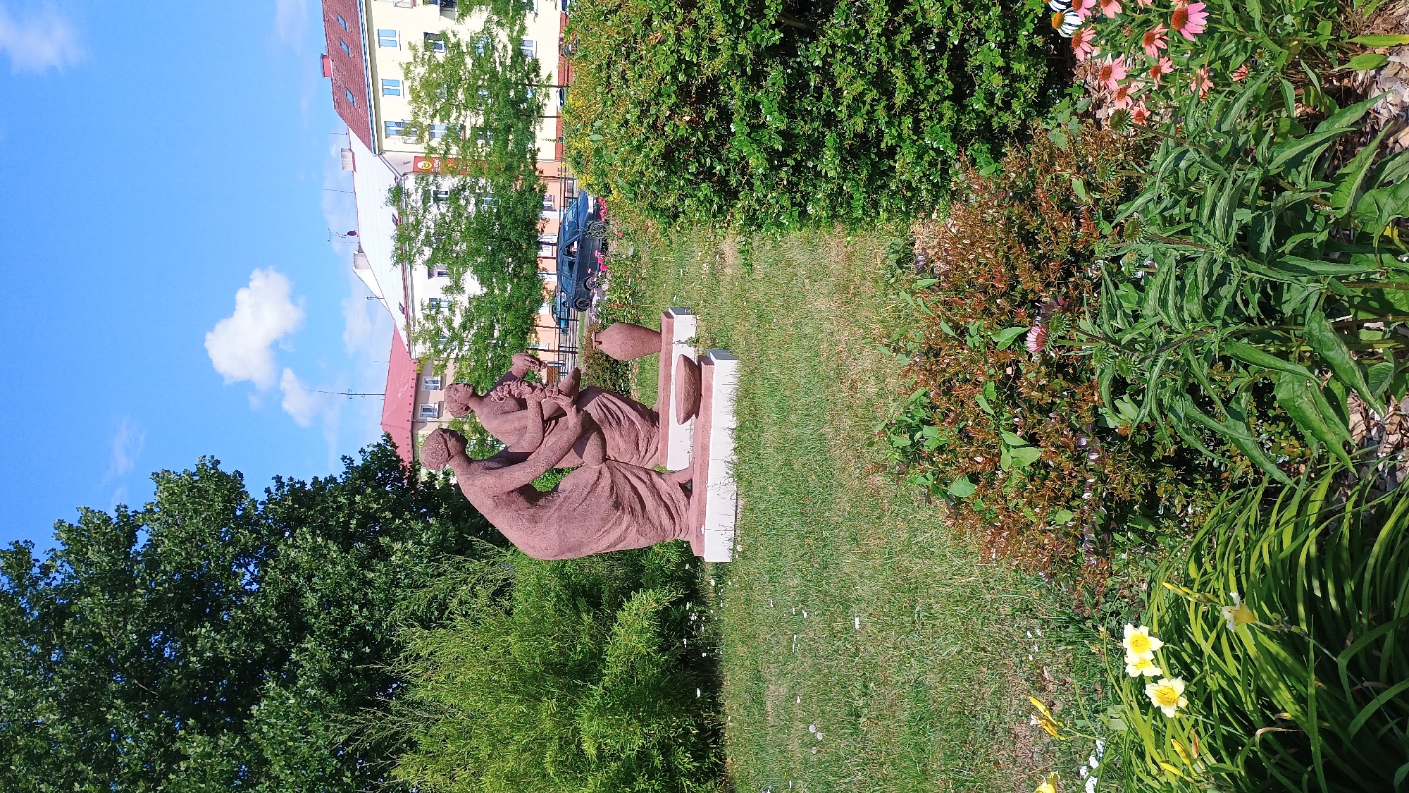 The image shows a reddish-brown sculpture of two women in flowing robes in a grassy park. The woman on the left is leaning over, and the woman on the right is holding a bundle of flowers or grapes. There is a large clay pot and a bowl in front of the sculpture. The sculpture is on a concrete base. Behind the sculpture, there is a large green bush, a line of trees, and buildings. In the foreground, there are various plants and flowers, including yellow lilies and pink coneflowers. The sky is blue with some white clouds. This sculpture is located in Sokolov, Czechia.