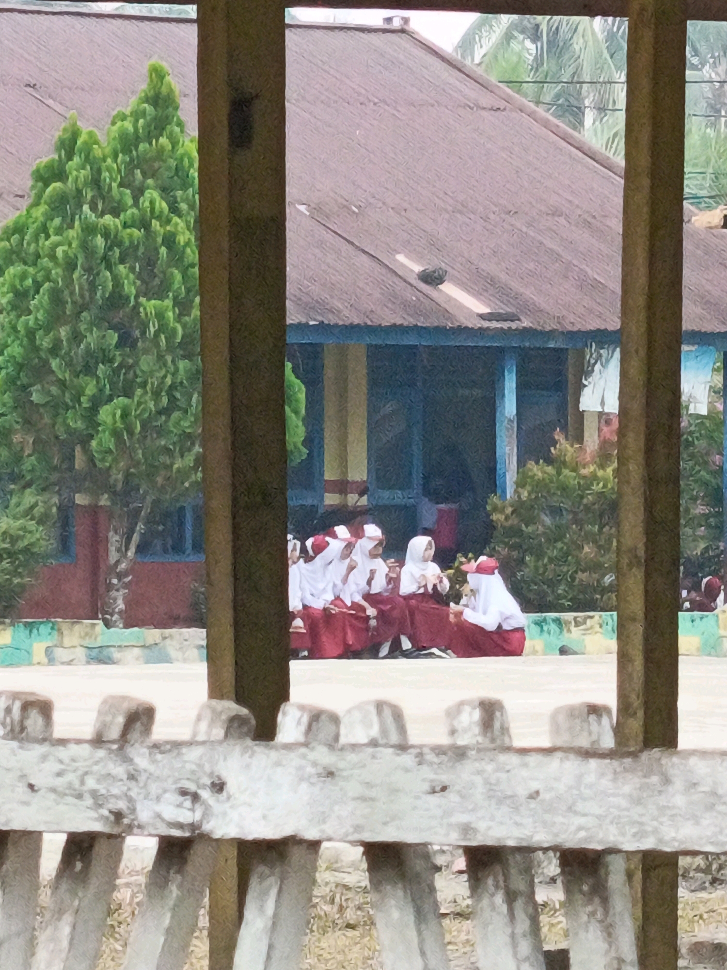 In Megang Sakti II, Indonesia, a group of students, likely attending a school, are visible. They are dressed in red skirts and white tops, with some wearing headscarves, suggesting a religious school or a uniformed event. They are seated on a paved area in front of a building with a corrugated iron roof. To the left, a large green tree stands out. In the foreground, a wooden fence partially obscures the view. The overall scene suggests a casual gathering or waiting period.