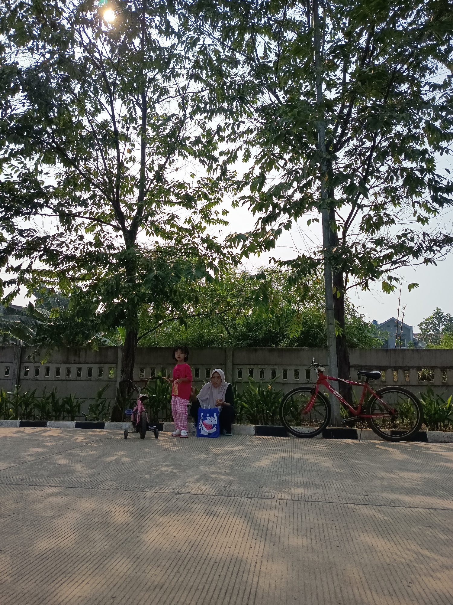 In a park setting in Bekasi, Indonesia, two individuals, a young girl and an adult woman, are positioned near a row of trees and a concrete fence. The young girl, wearing a pink t-shirt and plaid pants, stands beside a pink tricycle. The adult woman, wearing a hijab and dark clothing, is seated on the ground, next to a blue shopping bag with a cartoon character. A red bicycle is parked to the right of the woman. The scene is illuminated by sunlight filtering through the trees, casting shadows on the paved ground. The environment appears to be an outdoor recreational area with lush greenery and a clear sky.