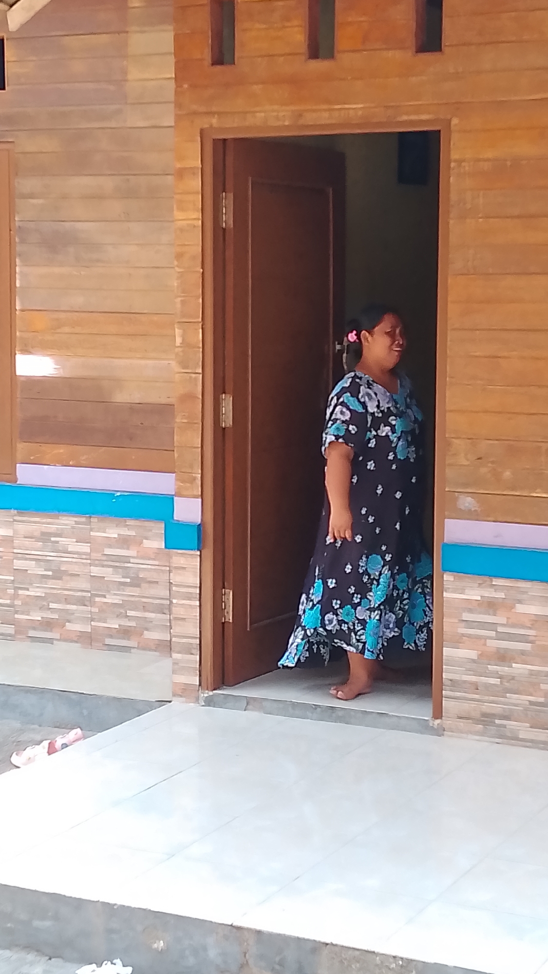 A woman stands in the doorway of a wooden house in Karanglangu, Indonesia. She is wearing a dark, floral-patterned dress with blue and white flowers. Her hair is pulled back with a pink accessory. The house has a wooden exterior with horizontal planks and decorative colorful bands below the windows. The ground in front of the house is tiled.