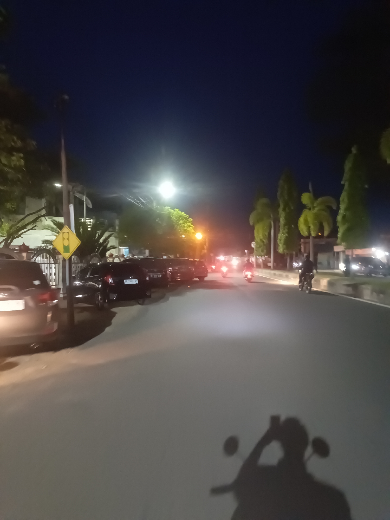 The image depicts a nighttime scene on a paved road, likely in Lhokseumawe, Indonesia. The sky is dark blue, suggesting it is either early evening or late night. Streetlights illuminate the scene, casting a warm glow on the road and surrounding objects.
Several parked cars are visible along the left side of the road. One car in the foreground on the left is dark and partially cut off by the frame. Further down the line, a dark SUV with a license plate reading "BK 1004 QI" is parked. Behind it, more vehicles are lined up.
On the right side of the road, a motorcycle rider is visible, casting a shadow on the road. The rider is silhouetted against the bright headlights of oncoming traffic. Other motorcycles with illuminated headlights can be seen further down the road, indicating ongoing activity. Palm trees and other foliage are scattered along the roadside, partially illuminated by the artificial lights.
A traffic signal sign, a yellow diamond with a black border and a depiction of a traffic light, is mounted on a pole on the left side of the road, near the parked cars. The overall atmosphere appears calm, with the activity of vehicles suggesting a typical urban or suburban environment. The lighting conditions create a somewhat moody ambiance. No specific weather conditions are discernible, but the clear sky indicates fair weather. The scene captures a moment of everyday life at night, with people commuting and vehicles navigating the roads.