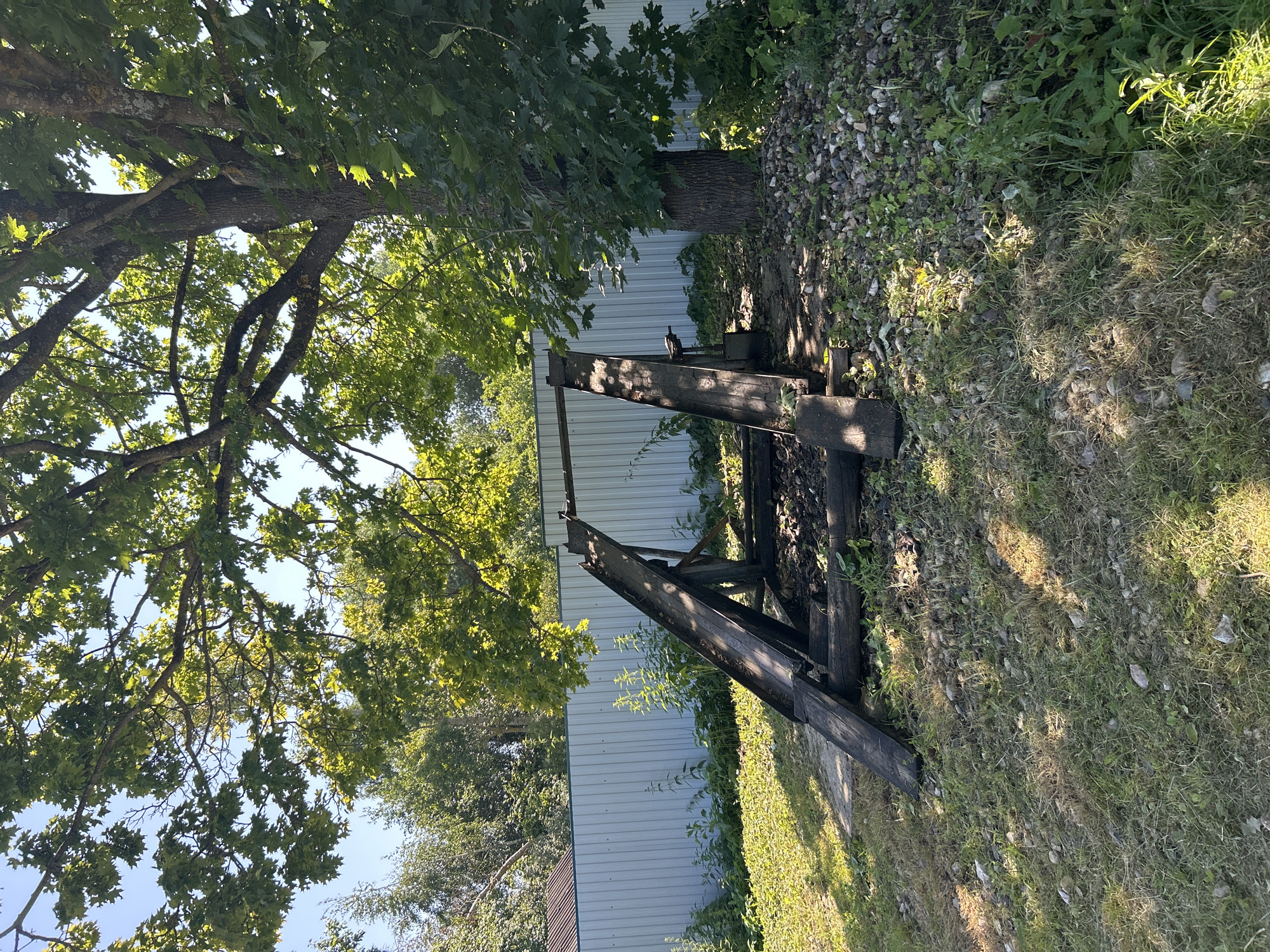 The image depicts an outdoor scene with a wooden structure that appears to be a ramp or part of a small bridge, constructed from weathered timber. The structure is situated on a grassy area with scattered small stones and debris. Behind the wooden structure is a light-colored, corrugated metal fence that runs horizontally across the scene. To the left of the fence, there are some trees and vegetation. A large tree with green foliage is prominent on the right side of the image, its branches partially obscuring the sky. The lighting suggests a sunny day, with shadows cast on the ground. There are no people or animals visible in the image. The location is identified as the city of Kalitsino, Russia.