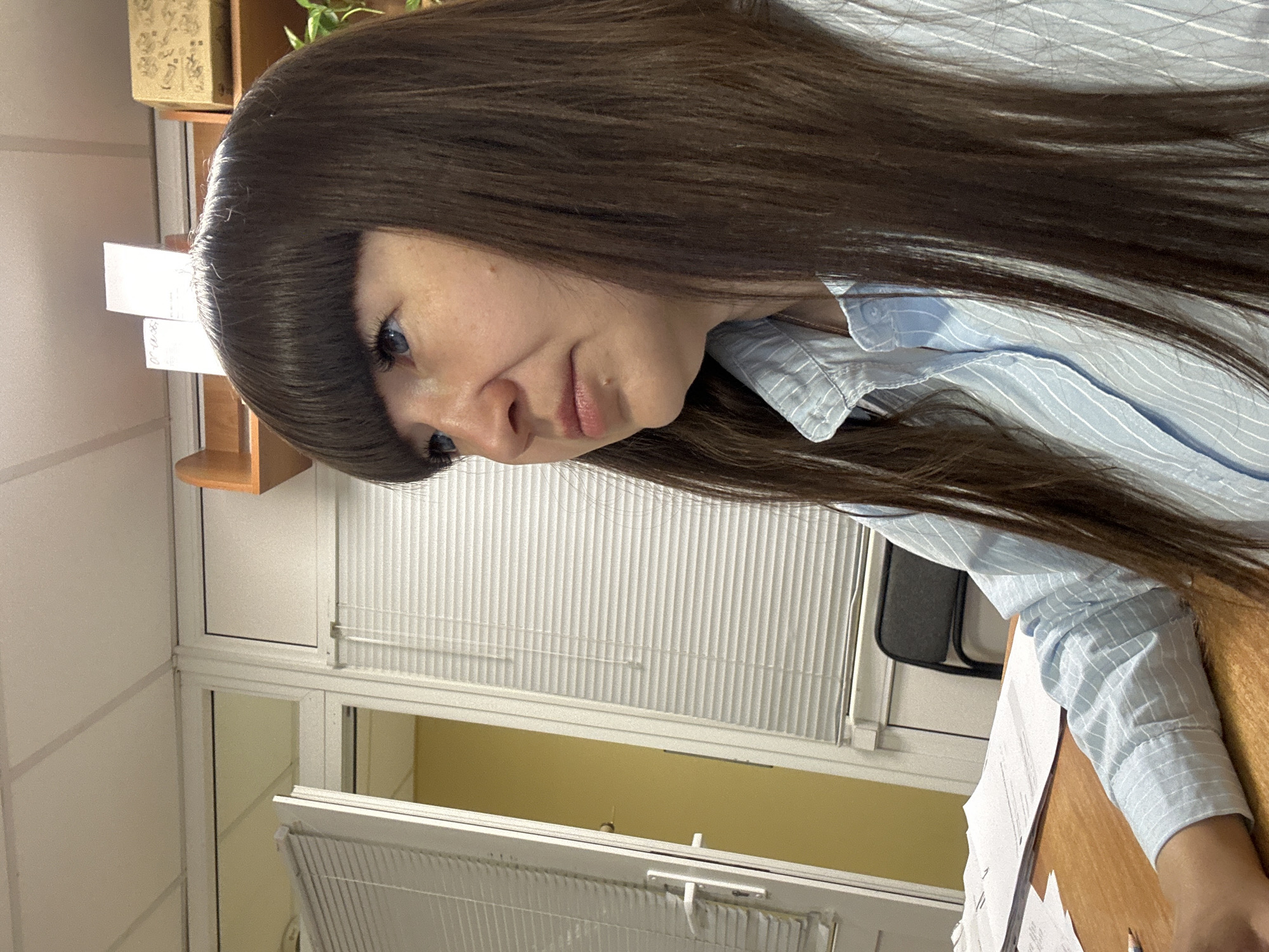 A medium shot angled downwards depicts a woman with long, brown hair and blue eyes. She wears a light blue striped shirt and sits at a wooden desk with papers on it. Background elements include a window with white blinds, a beige wall, and an office chair. The location is possibly an office setting in the city of Chaikovsky, Russia.