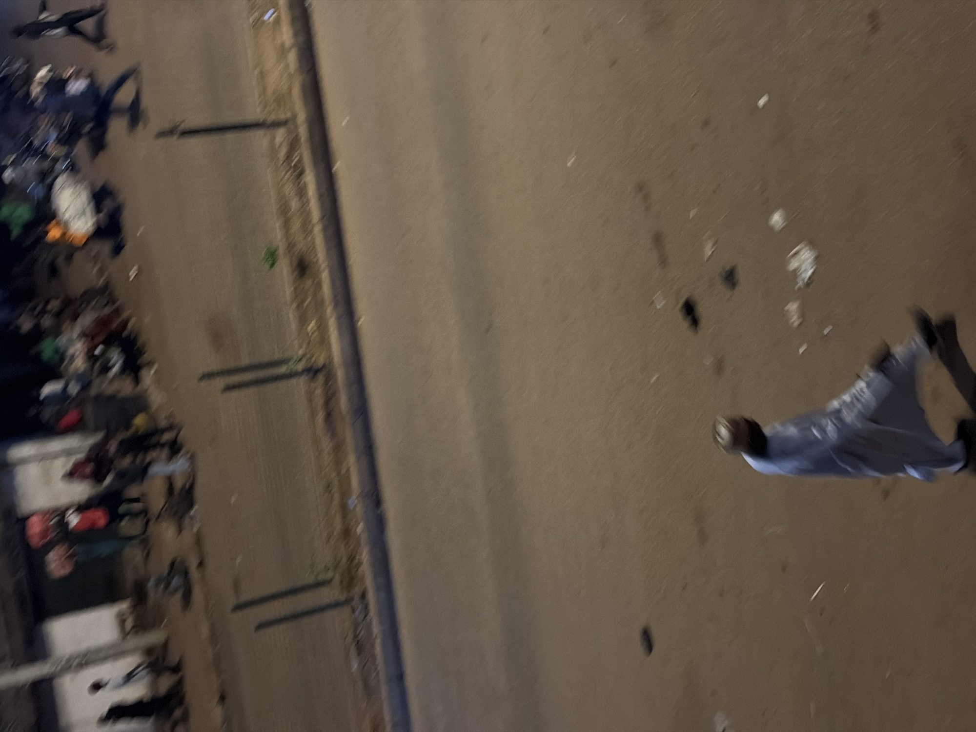 The image shows a street scene in Bukuru, Nigeria at night, illuminated by artificial light. In the foreground, a person wearing a light-colored, long garment and a face covering walks across the dusty street. Further up the street, a group of people are visible in front of buildings. Some individuals appear to be carrying items. The street is lined with a curb and a fence made of vertical metal posts. The ground is uneven and appears to have some debris scattered across it. The overall impression is of a busy urban environment at dusk or nighttime.