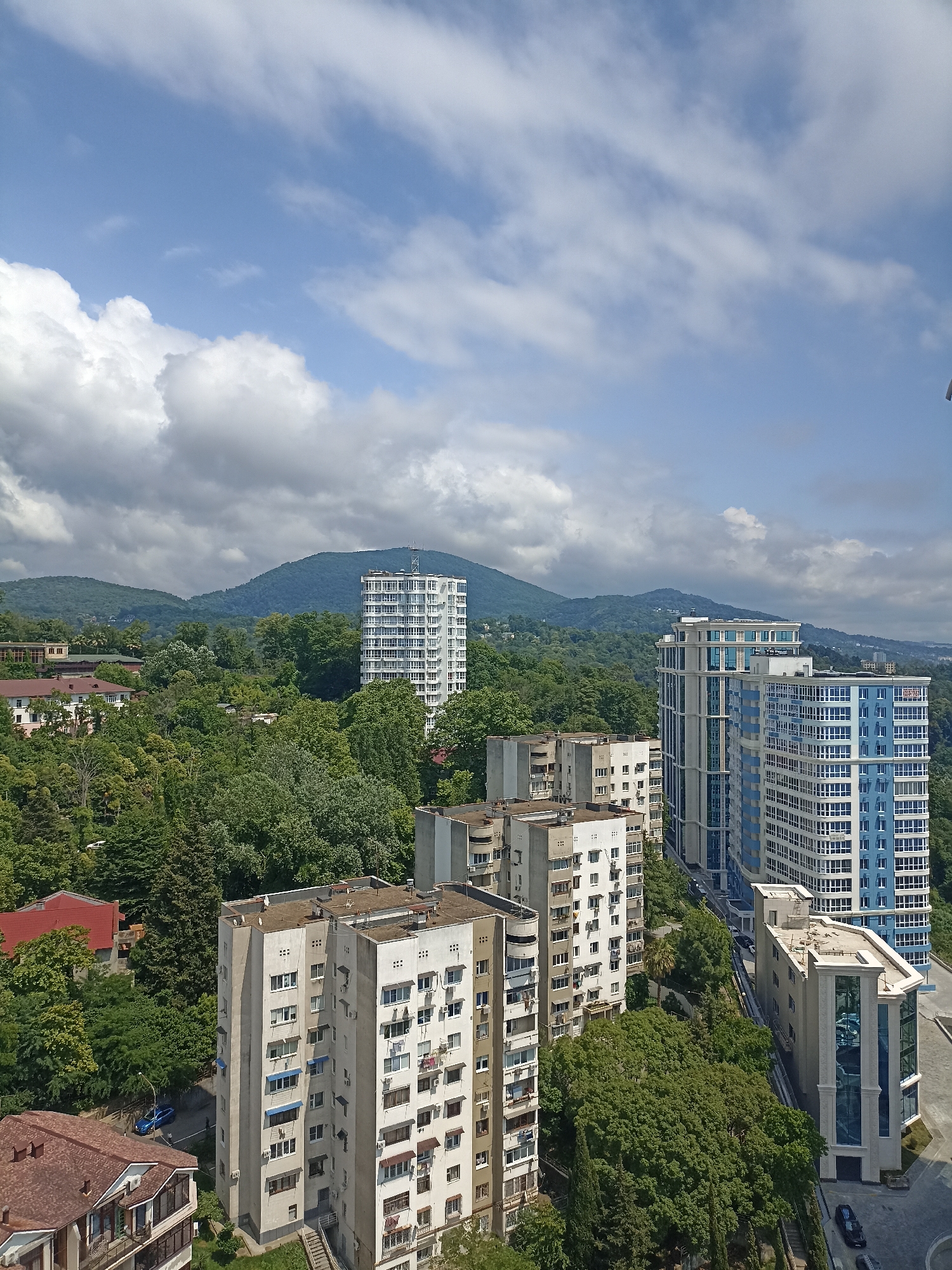The image captures a cityscape in Sochi, Russia, characterized by a blend of residential buildings and lush natural surroundings. In the foreground, several apartment buildings of varying heights and architectural styles are visible. One prominent building is a tall, white structure with numerous windows and balconies, set against a backdrop of green trees. Adjacent to it, a cluster of older, beige-colored apartment blocks stands, their facades marked by many windows and air conditioning units. Further into the scene, modern high-rise buildings with blue and white facades dominate the skyline, their reflective windows creating a striking contrast with the natural landscape.
The middle ground is densely populated with trees, creating a vibrant green canopy that partially obscures the lower parts of the buildings. This abundant vegetation suggests a city integrated with nature, common in coastal and mountainous regions like Sochi. In the distance, rolling hills covered in dense forest rise, their slopes softened by the haze of a warm day. The sky above is a mix of clear blue and scattered white clouds, some of which are large and fluffy, casting shadows on the landscape. The sunlight indicates it is daytime. A blue car is parked on a street visible between the buildings and trees. The overall impression is one of a developed urban area nestled within a picturesque natural setting, characteristic of Sochi's topography.