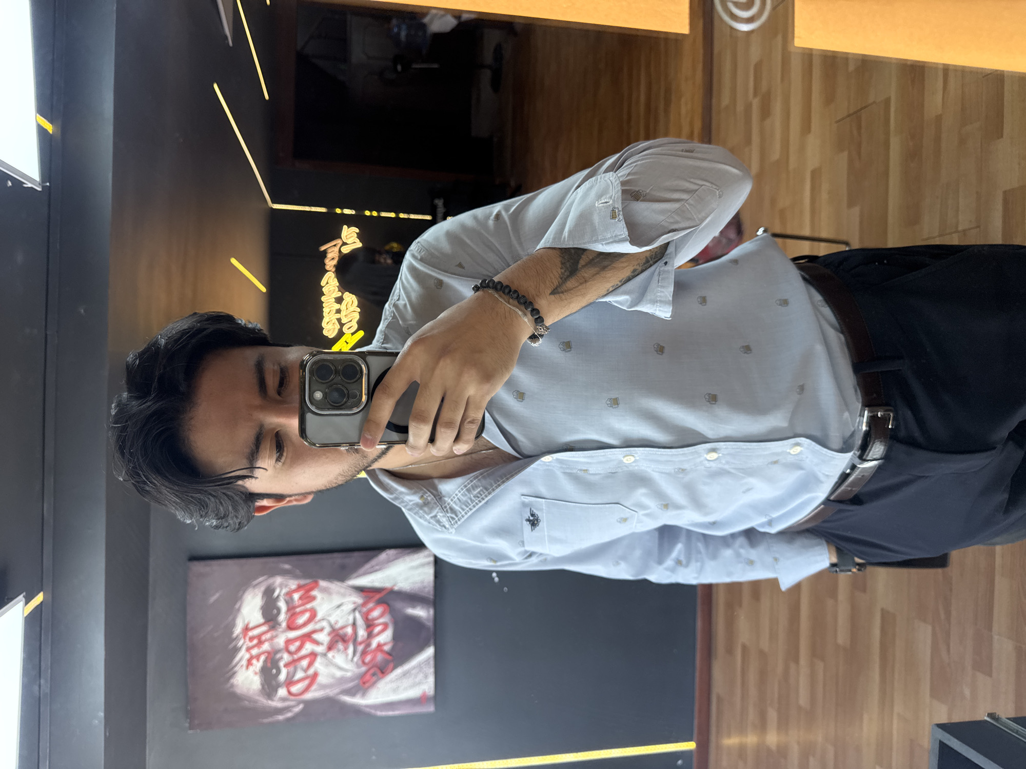 A young man is captured in a mirror selfie, holding a grey iPhone with multiple cameras that partially obscures his face. He has dark hair and light stubble. He is wearing a light blue, short-sleeved button-up shirt adorned with small beer mug patterns and a dark emblem on the left chest pocket. His attire includes dark trousers and a brown belt. Accessories consist of a dark beaded bracelet on his right wrist and a thin chain necklace. A tattoo is visible on his right arm.
The setting appears to be a trendy indoor commercial space, potentially a bar or restaurant in Puebla, Mexico. The background features a dark wall to the left, a wooden floor to the right, and wooden paneling at the top right. A prominent painting on the dark wall depicts a stylized face, resembling the Joker, with the red text "WHY SO SERIOUS?" written across it. Above the painting, a yellow neon sign displays script text that reads "Tapas bistro" followed by additional words, possibly "Puebla". Another bright, white light fixture is visible on the far left. The artificial lighting suggests an indoor setting where the time of day is not a primary factor.