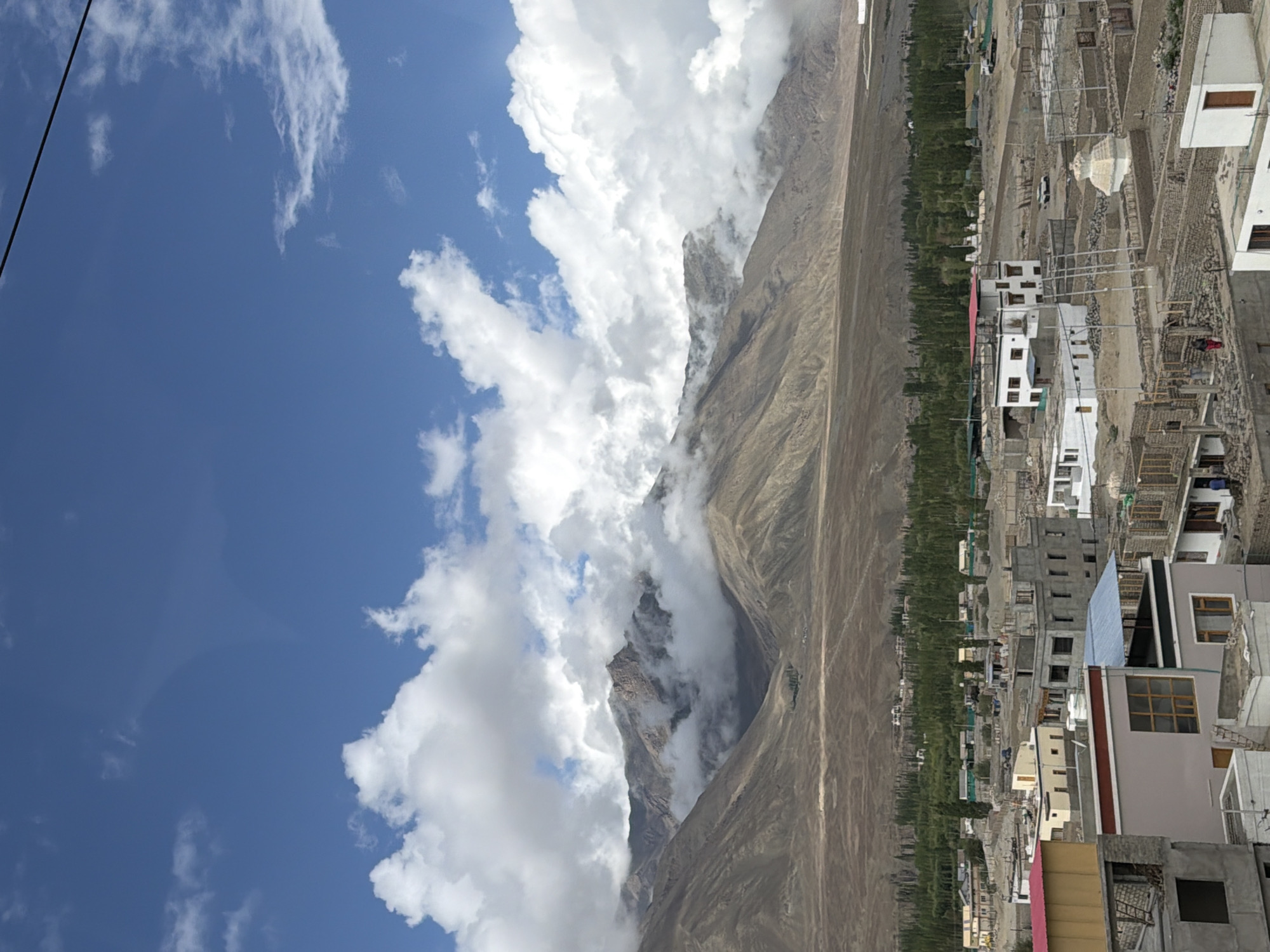 This is a vertical, eye-level, full shot of the Spituk region of Leh, India, on a bright day with scattered clouds. The background is dominated by rugged, brown mountains that are partially obscured by thick, white clouds. In the midground, a lush green forest stretches across the landscape, creating a stark contrast with the arid mountains. Below the forest, a village is visible, characterized by numerous buildings of varying sizes and architectural styles. Some buildings are constructed with concrete, while others appear to be made of mud bricks. Construction activity is evident in some areas, with unfinished structures and scaffolding. In the foreground, several buildings are more detailed, showing their facades, windows, and roofs. A prominent white building with a red roof stands out. A small stupa-like structure is also visible in the village. A few people, appearing as small figures, are scattered throughout the scene, engaged in their daily activities. The sky is a vibrant blue, with the white clouds casting shadows on the mountains. A black electrical wire is visible crossing the top of the frame.