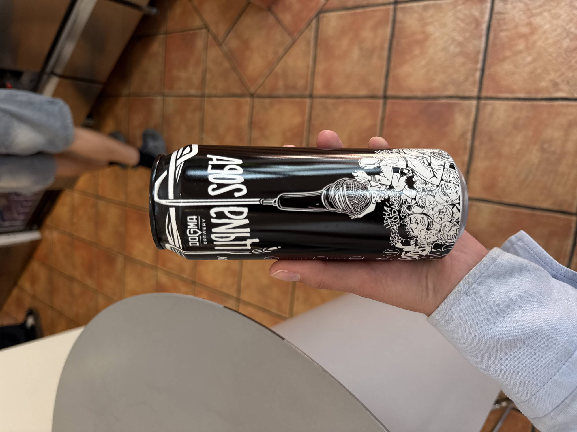 A person is holding a black and white "Dogma Brewery" craft beer can labeled "Sons Enbitch" featuring a microphone graphic. The photo was taken in Belgrade, Serbia, as the brand is from that city. The person is sitting at a table, wearing a light blue shirt. The background includes a tiled floor and part of a kitchen or serving area. Someone sitting nearby is wearing sweatpants.