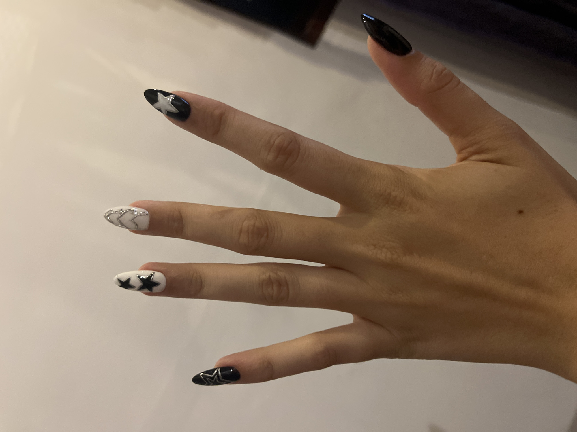 A person's left hand is shown with stiletto-shaped nails. The nails are decorated with black and white polish and silver star accents. The background is a plain, light-colored wall. The image appears to have been taken indoors.