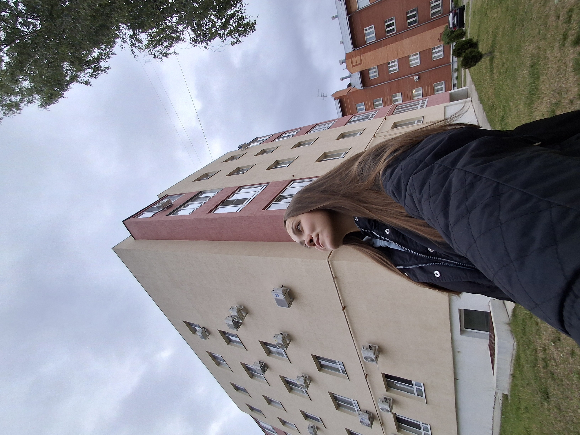 The image, captured in Chaikovsky, Russia, presents a low-angle perspective of an urban residential scene under an overcast sky. In the immediate foreground, a young woman with long brown hair is visible from the chest up, looking upward and slightly to her right with a subtle smile. She wears a dark, possibly quilted, jacket over a light blue garment. Her right arm extends towards the camera, suggesting she is holding the device.
Dominating the left and central portions of the background is a multi-story residential building. Its primary visible facade is cream-colored, with a corner section displaying a reddish-brown hue. The cream facade features numerous evenly spaced rectangular windows with white frames, below or beside which various outdoor air conditioning units are mounted. A light-colored pipe or conduit runs horizontally along the lower part of this wall. At the ground level, a small basement window and a dark grate are visible.
To the right, behind the first building, another multi-story residential structure is partially visible. This building is constructed primarily of reddish-brown brick and also features multiple rectangular windows and distinct rooflines with an antenna.
The ground around the buildings is covered with green grass, showing some patches of dryness. In the far right, a large deciduous tree with green leaves is partially visible, extending into the upper corner of the frame. Several small, conical evergreen bushes are planted on the lawn between the buildings, and a dark-colored car is partially visible parked behind the background building. Overhead, thin utility wires are discernible against the uniform light grey, cloudy sky.