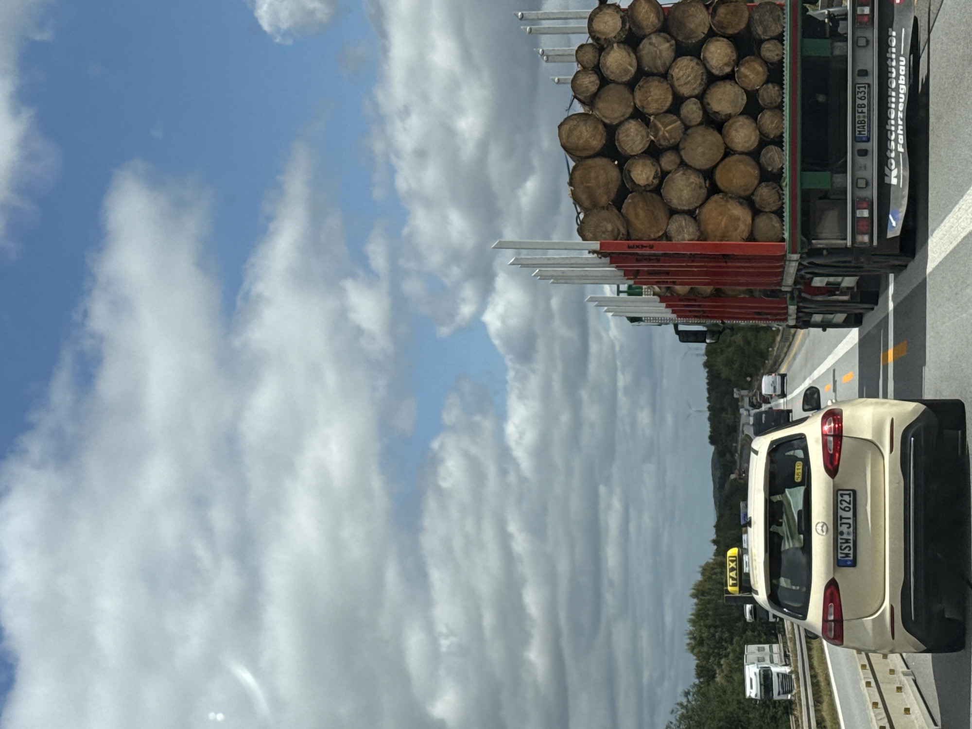 The image captures a view from inside a vehicle traveling on a highway, likely in Burkau - Porchow, Germany, given the context. The foreground features the rear of a yellow Mercedes-Benz taxi with the license plate "WSW JT 621". To the right, a large truck loaded with logs is prominently visible, with "Kotschenreuther Fahrzeugbau" inscribed on its rear. Other vehicles, including a white semi-truck and various cars, are present on the highway in the background. The sky is partly cloudy with a blue sky visible through the clouds. The environment appears to be a rural or semi-rural highway with trees lining the roadside.