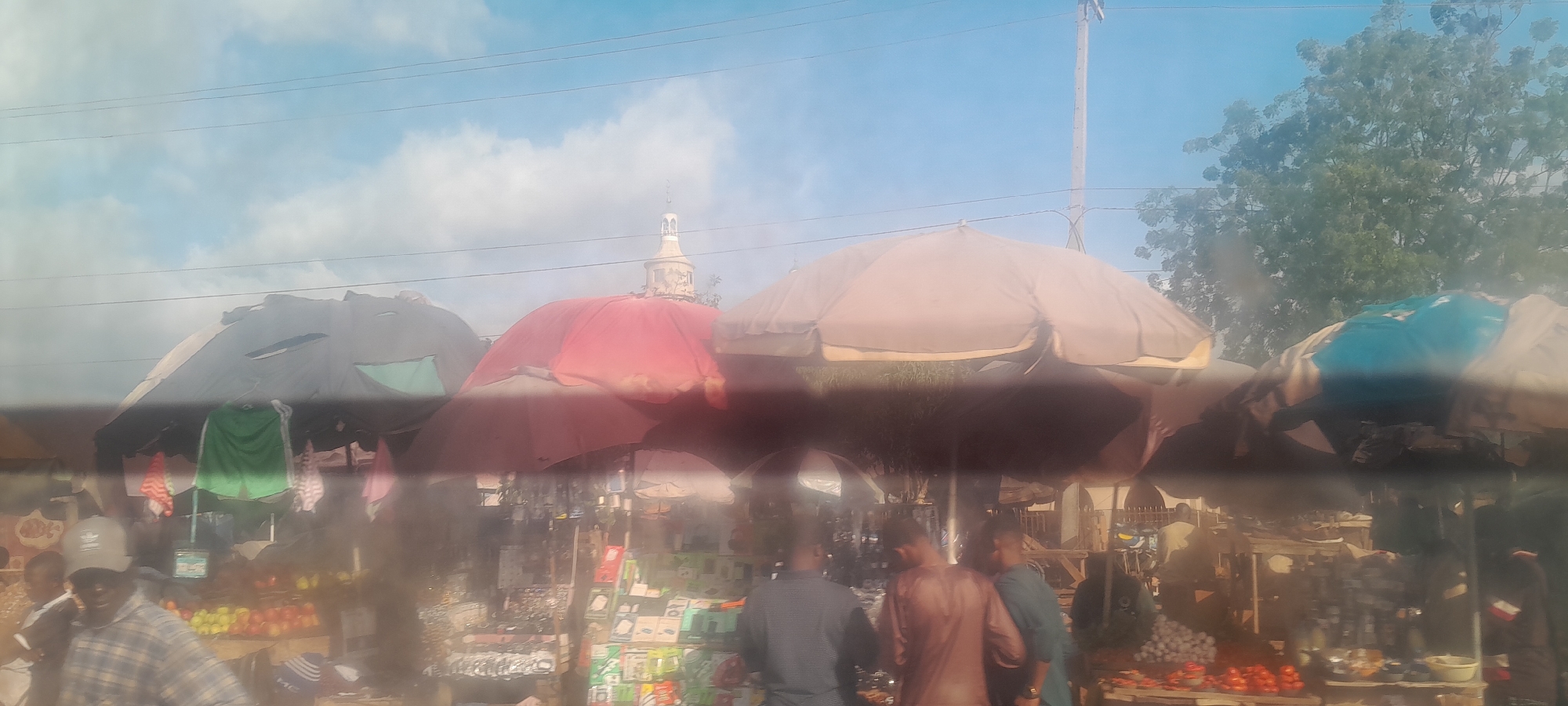 The image captures a bustling street market in Nyanya, Nigeria, under a clear, cloudy sky. Several large market stalls with colorful umbrellas, predominantly red, tan, and grey, provide shade. One stall features a prominent green garment hanging, possibly shorts or a shirt. In the background, a tower-like structure with an ornate top is visible, suggesting a landmark. A variety of goods are displayed at the stalls, including what appear to be fruits, such as tomatoes and yellow fruits, and various packaged items. Several individuals are present, engaged in shopping or selling activities. One man in the foreground wears a plaid shirt and a cap. The scene is filled with the vibrant energy of a local market.
