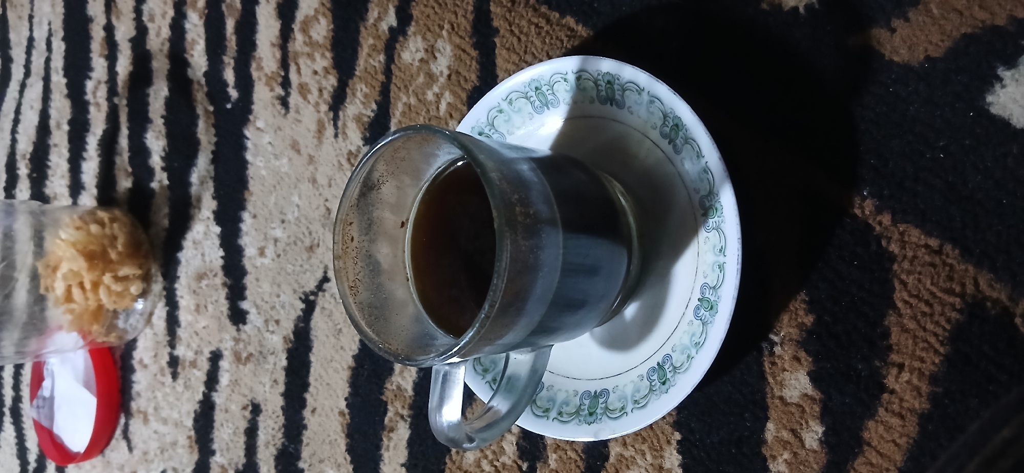 A clear glass mug filled with dark liquid, likely coffee, rests on a white saucer with a green patterned rim. The mug and saucer are positioned on a carpet with a black and tan striped pattern. In the background, to the upper left of the mug, a clear plastic container with a red lid is visible, containing a small pile of light brown, crunchy-looking food items. The scene appears to be indoors, possibly in a home in Demangan, Indonesia, given the context. There are no people or actions depicted.