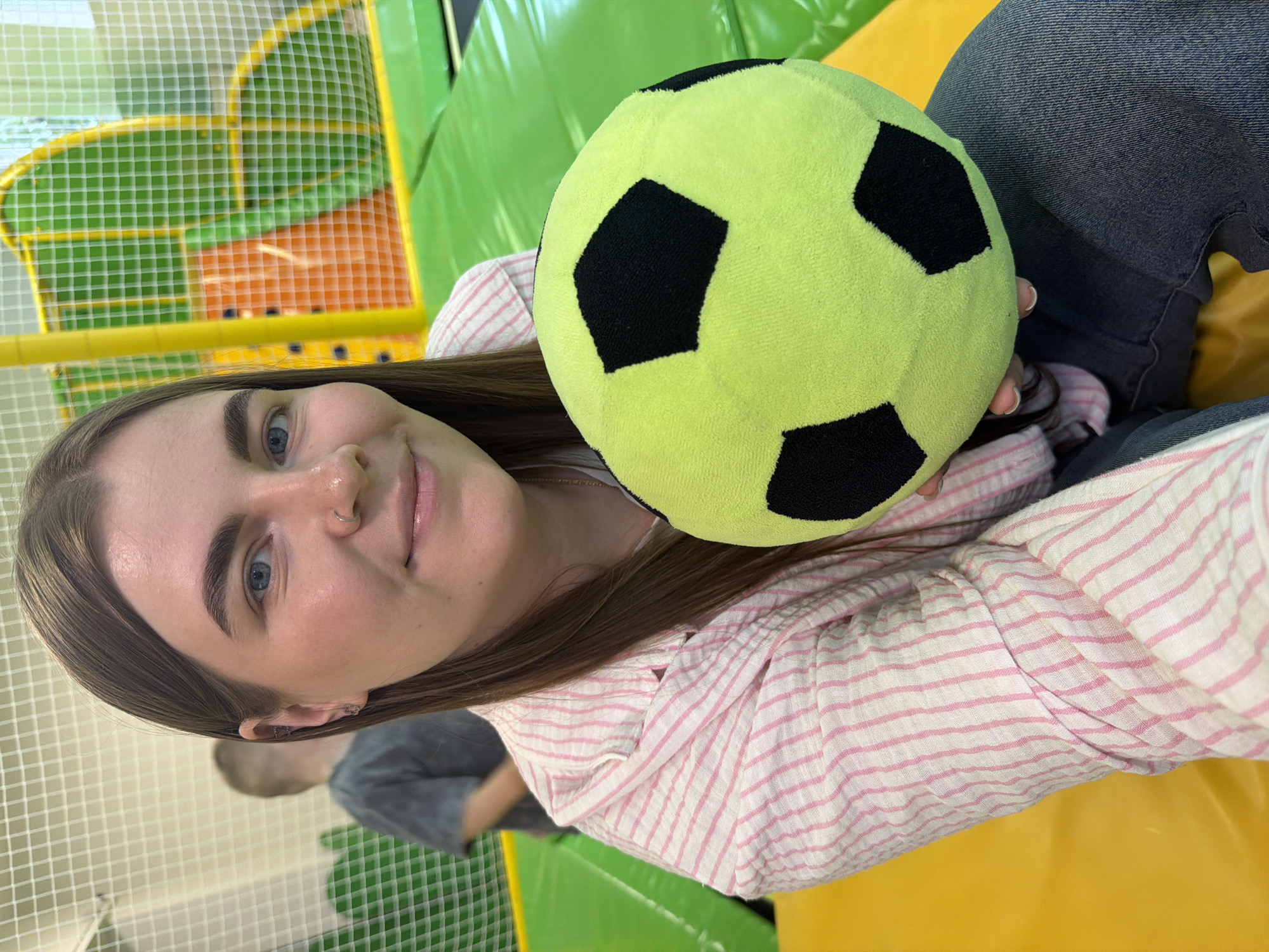 A woman holds a green and black soccer ball. She is wearing a white and pink striped top and blue jeans. The background is a children's play area, possibly in the city of Chaikovsky, Russia.