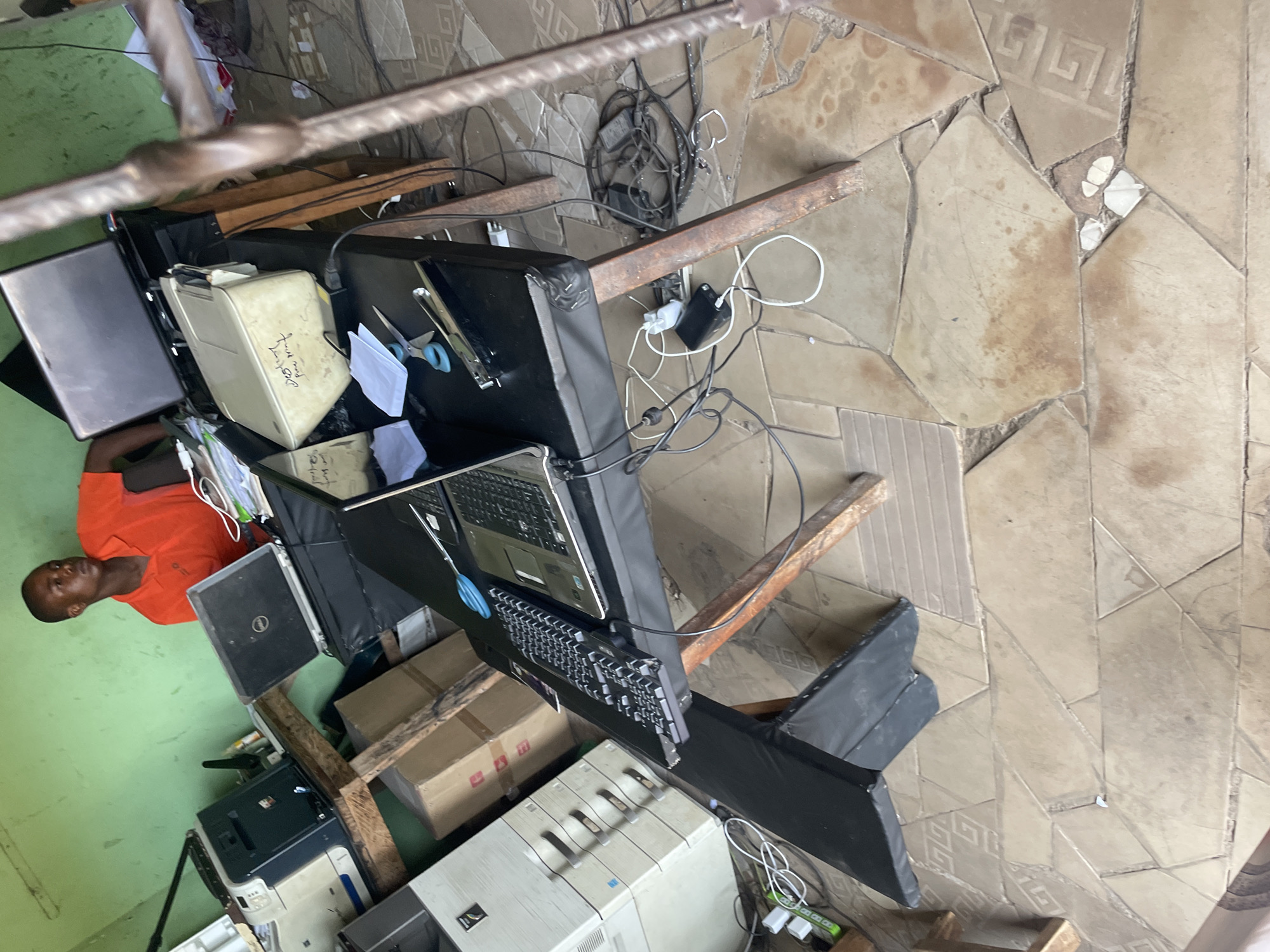 The image depicts an indoor workspace in Mararaba, Nigeria. A man wearing an orange shirt is seated at a wooden desk with multiple computers, printers, and electronic devices. Various power cords and cables are visible. The room has green walls, and the floor is tiled with geometric patterns. Boxes and other office supplies are present. The lighting is bright.